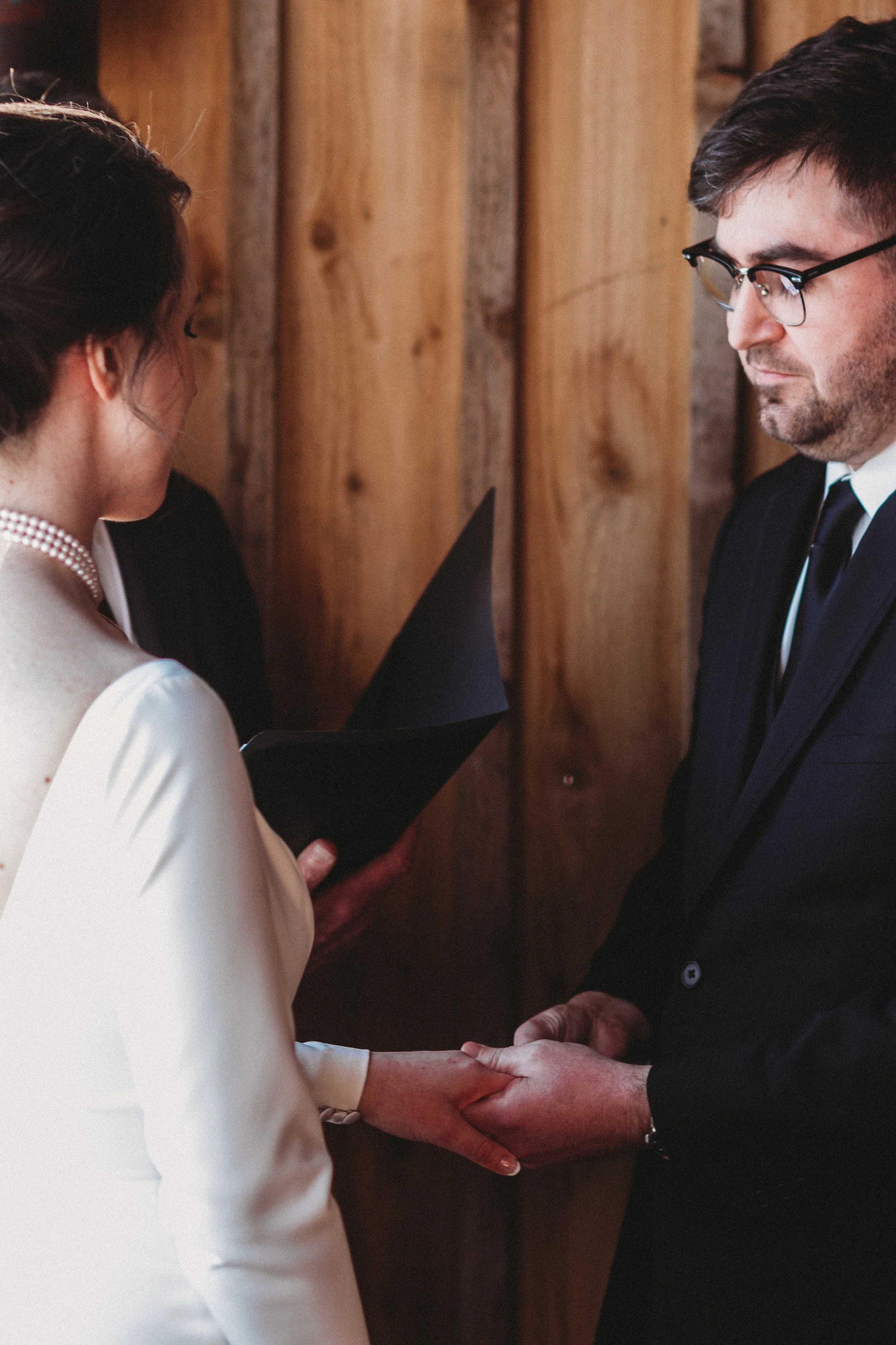 A couple holding hands during a wedding ceremony, with the bride reading vows and the groom looking at her, standing against a wooden background.