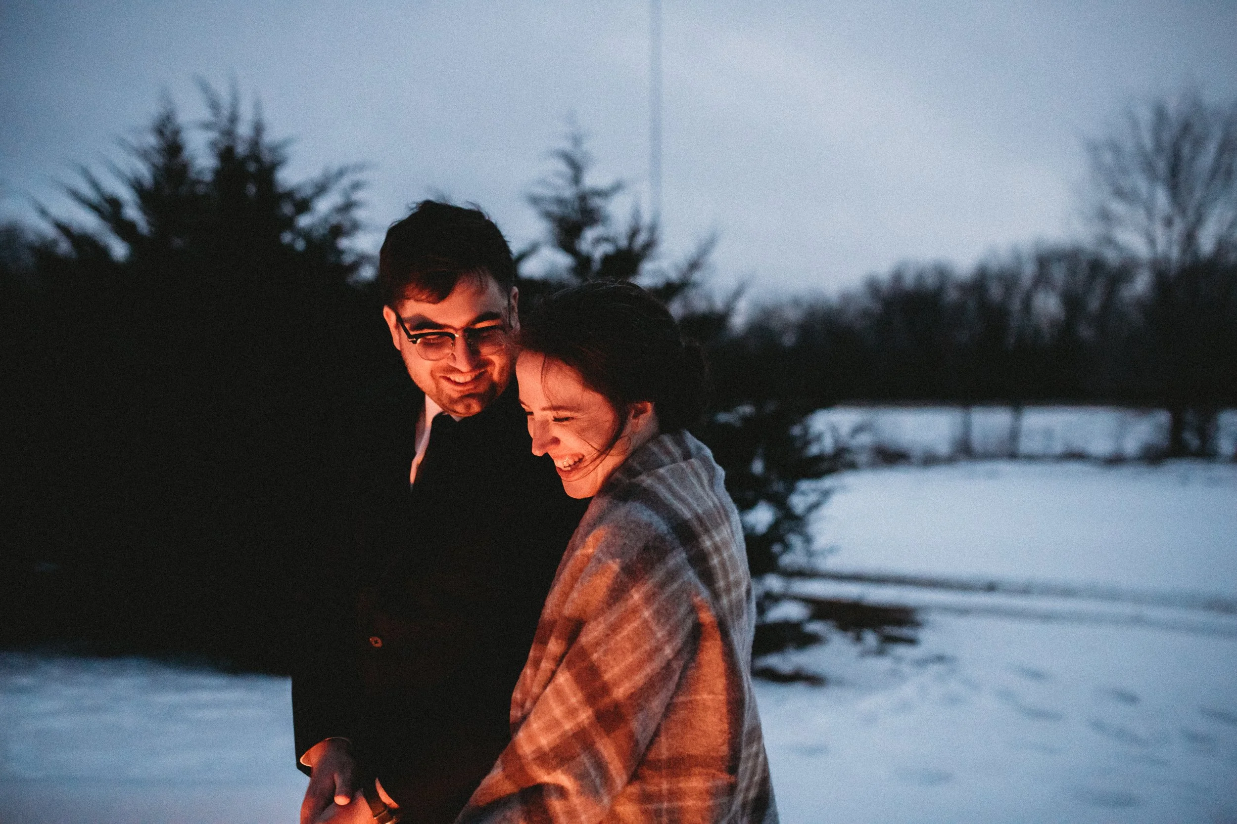 A man and woman smiling and laughing outdoors during winter, with snow on the ground and bare trees in the background, illuminated by a small fire or light