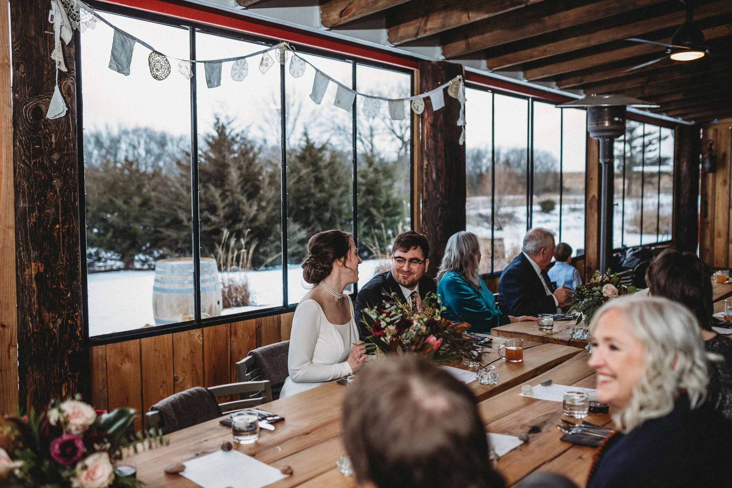 People gathered at a rustic indoor wedding reception or celebration, sitting at a long wooden table decorated with floral arrangements, with a large window offering a snowy outdoor view.