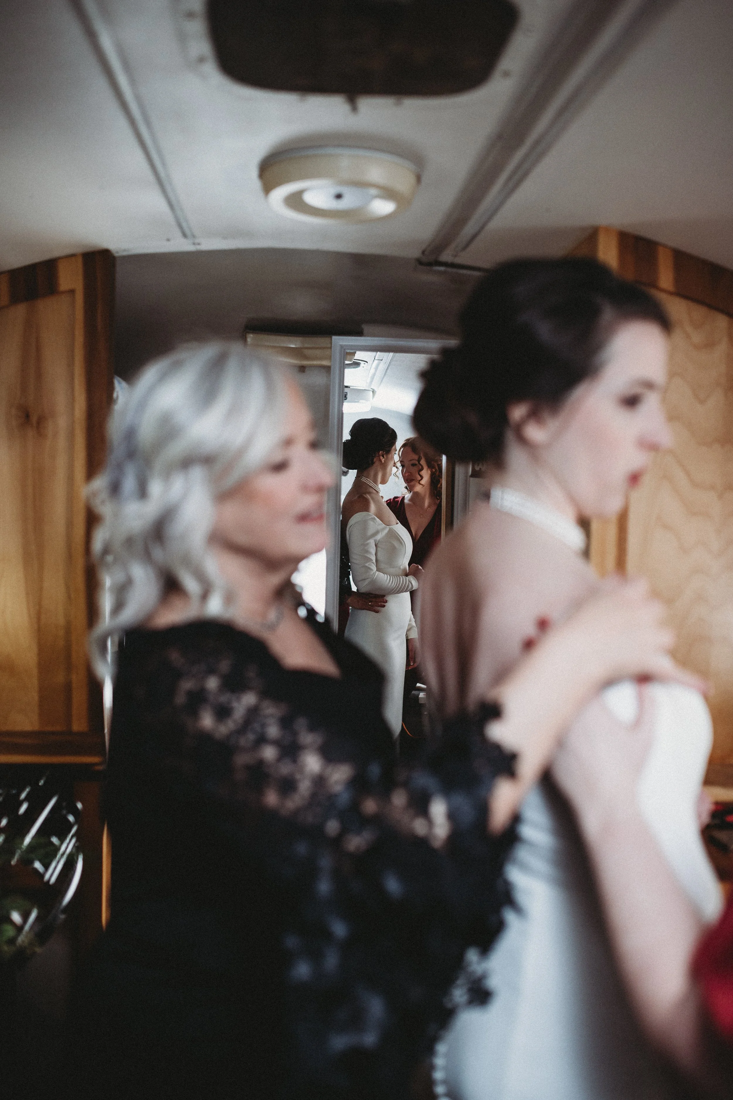 Women in elegant dresses preparing for a wedding, with two women in the background seen through a doorway.
