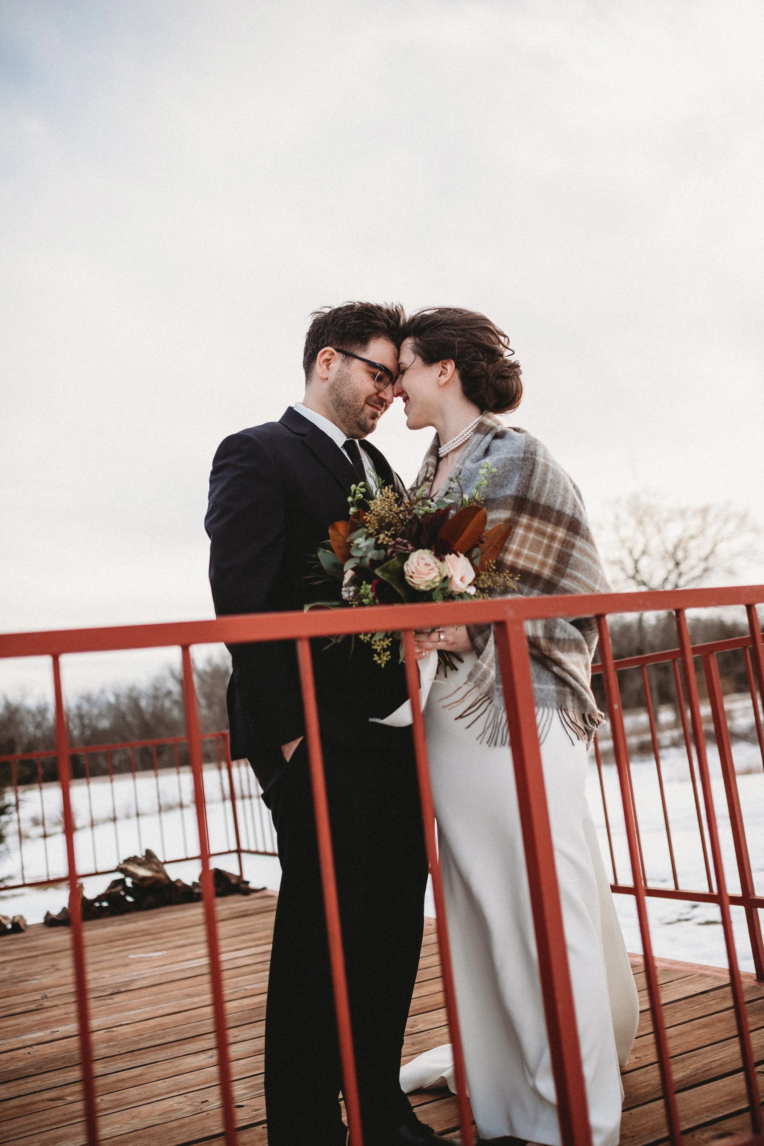 A couple dressed in formal wedding attire, standing on a wooden bridge outdoors, with the woman holding a bouquet of flowers, touching foreheads, and smiling softly. Burning Barrel Lecompton Kansas. 