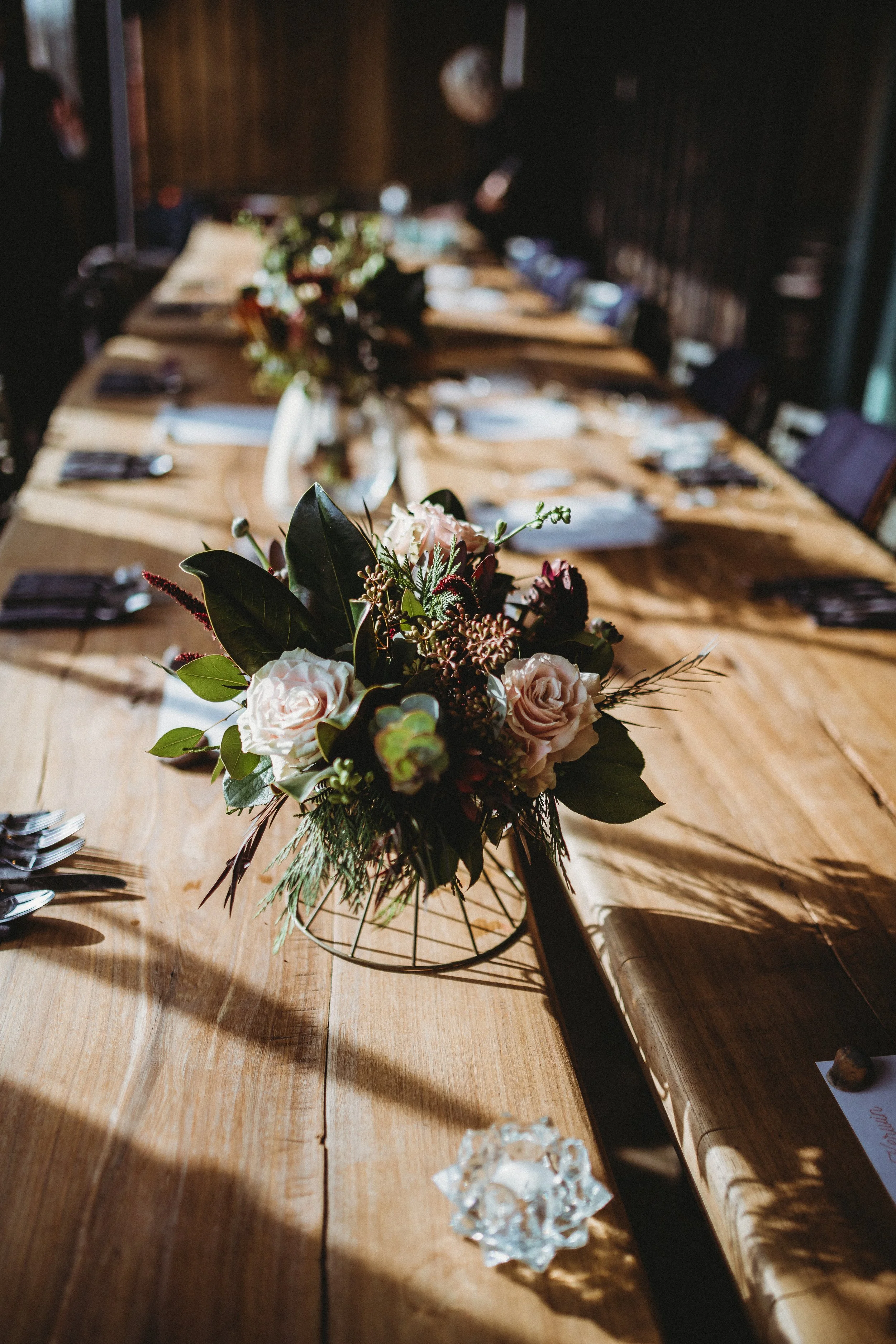 A floral centerpiece with pink roses and green foliage on a wooden dining table set for a meal.