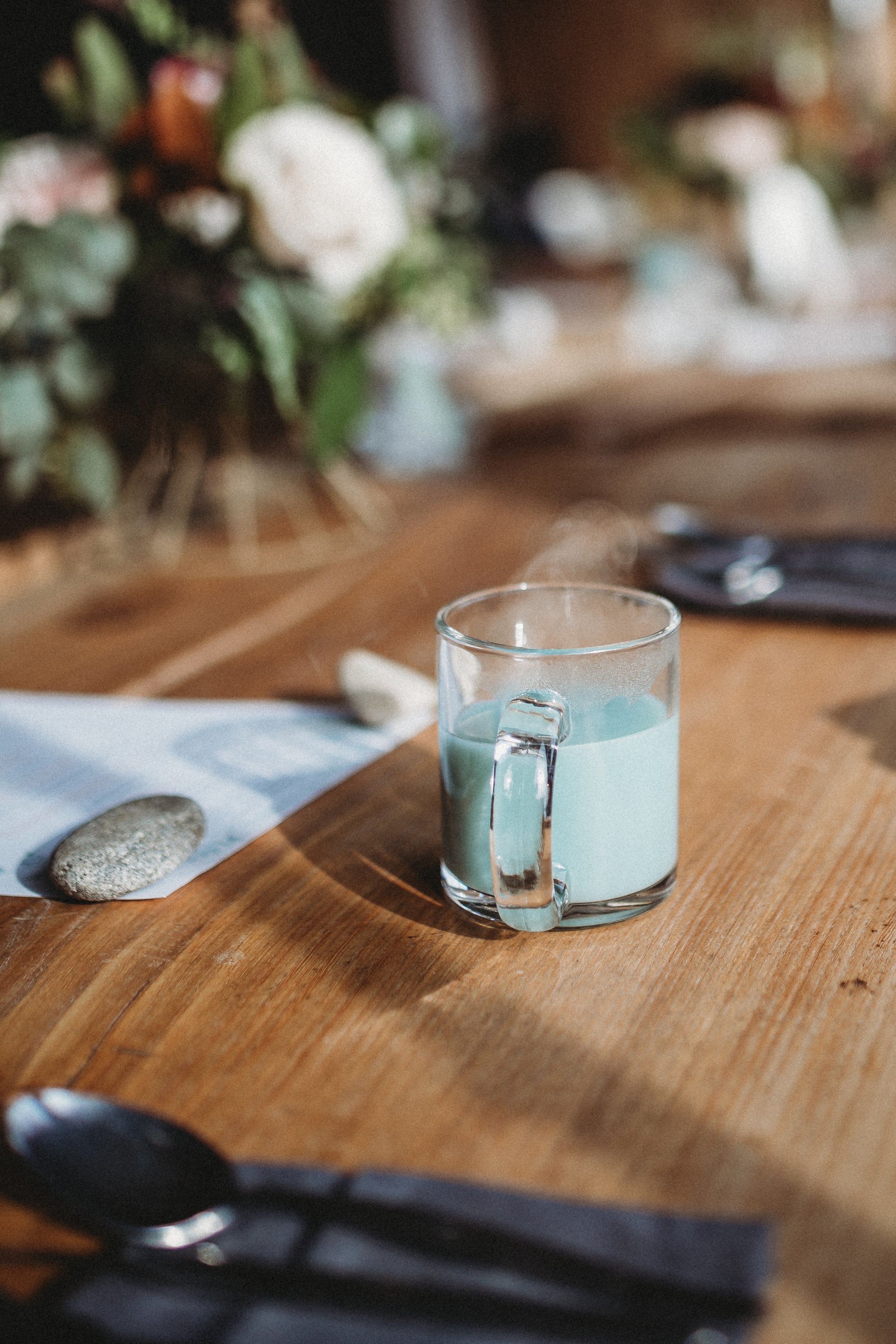 A steaming cup of coffee or tea on a wooden table with utensils and a floral centerpiece in the background.
