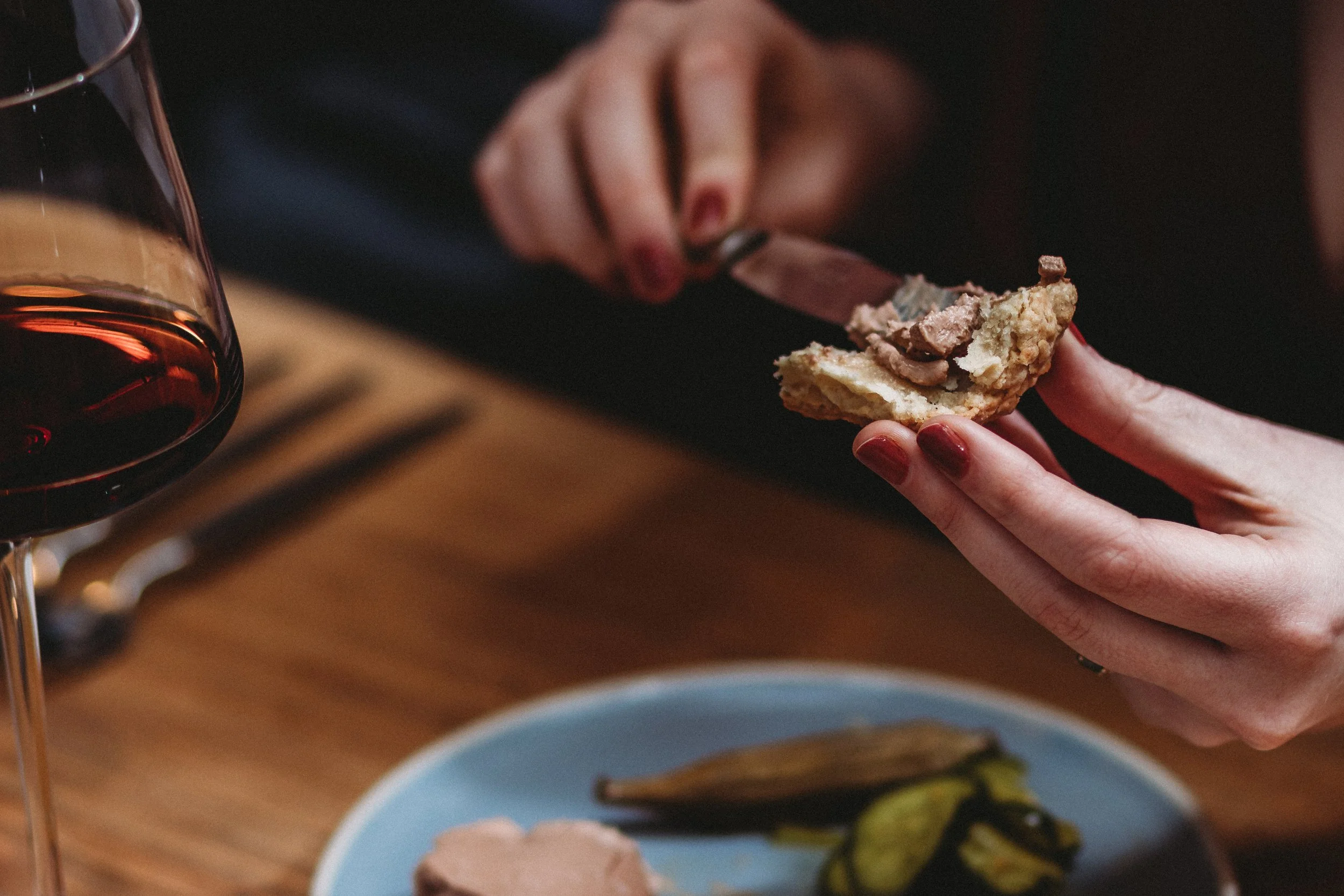 Person spreading butter on a slice of bread with a knife at a dining table, accompanied by a glass of red wine and some pickles on a blue plate.