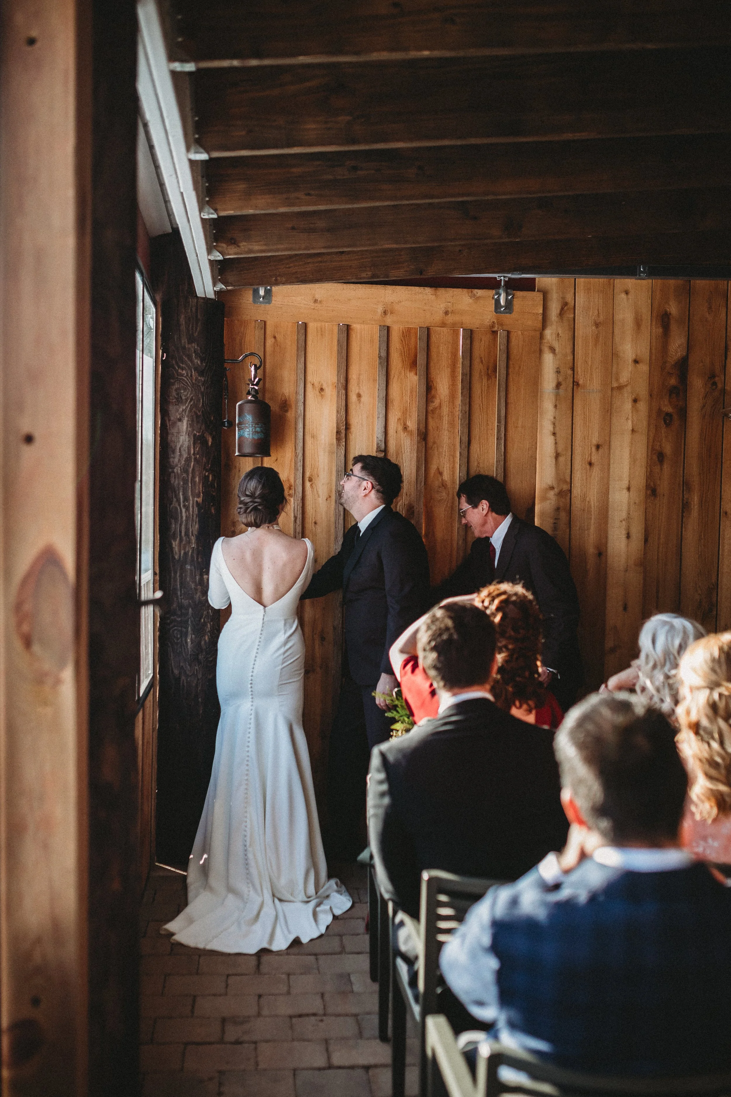 A bride and groom holding hands during a wedding ceremony, standing in front of a wooden wall, with seated guests observing.