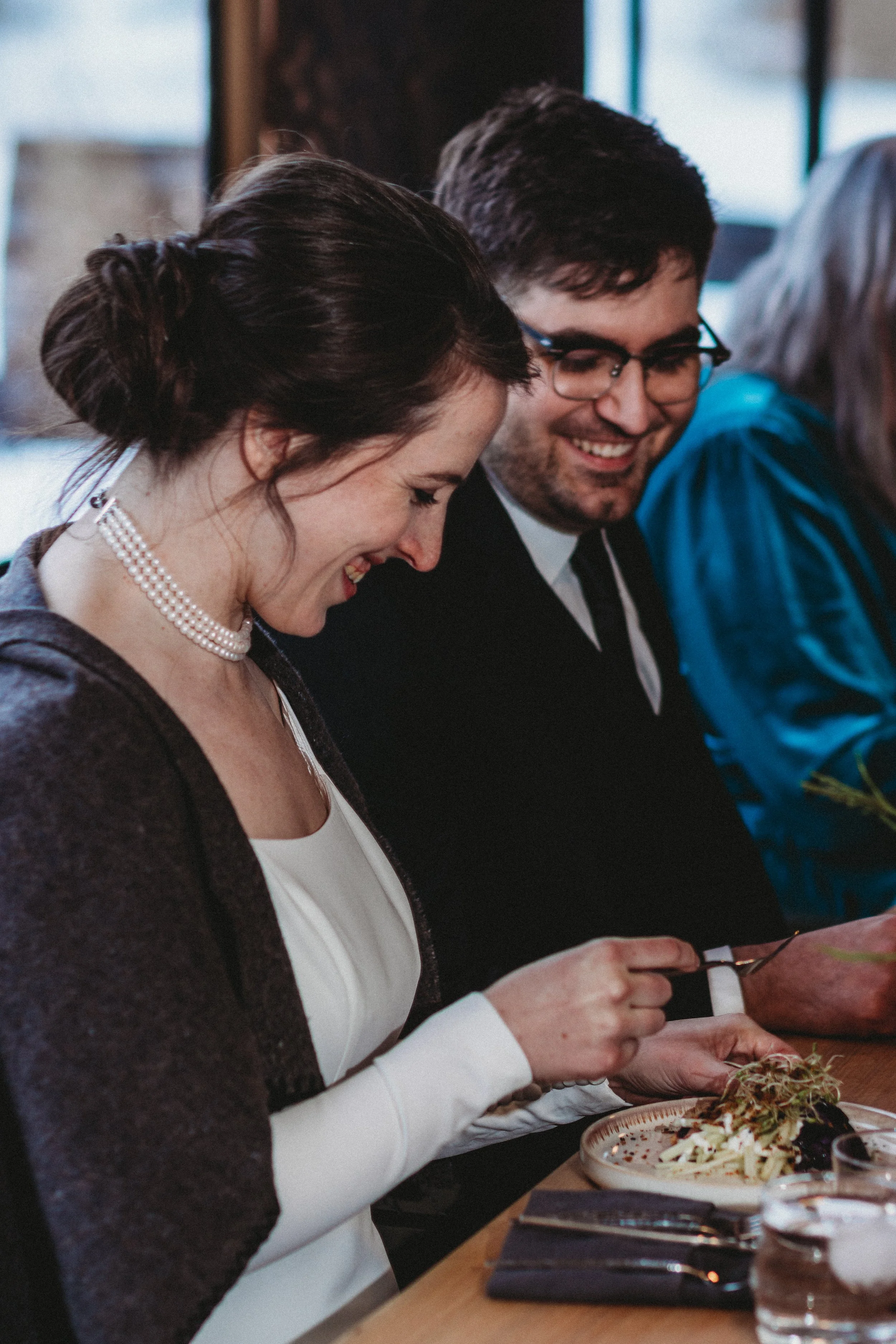 A woman and a man are sitting together at a dining table, smiling and looking at her plate of food, which appears to be pasta with garnishes.