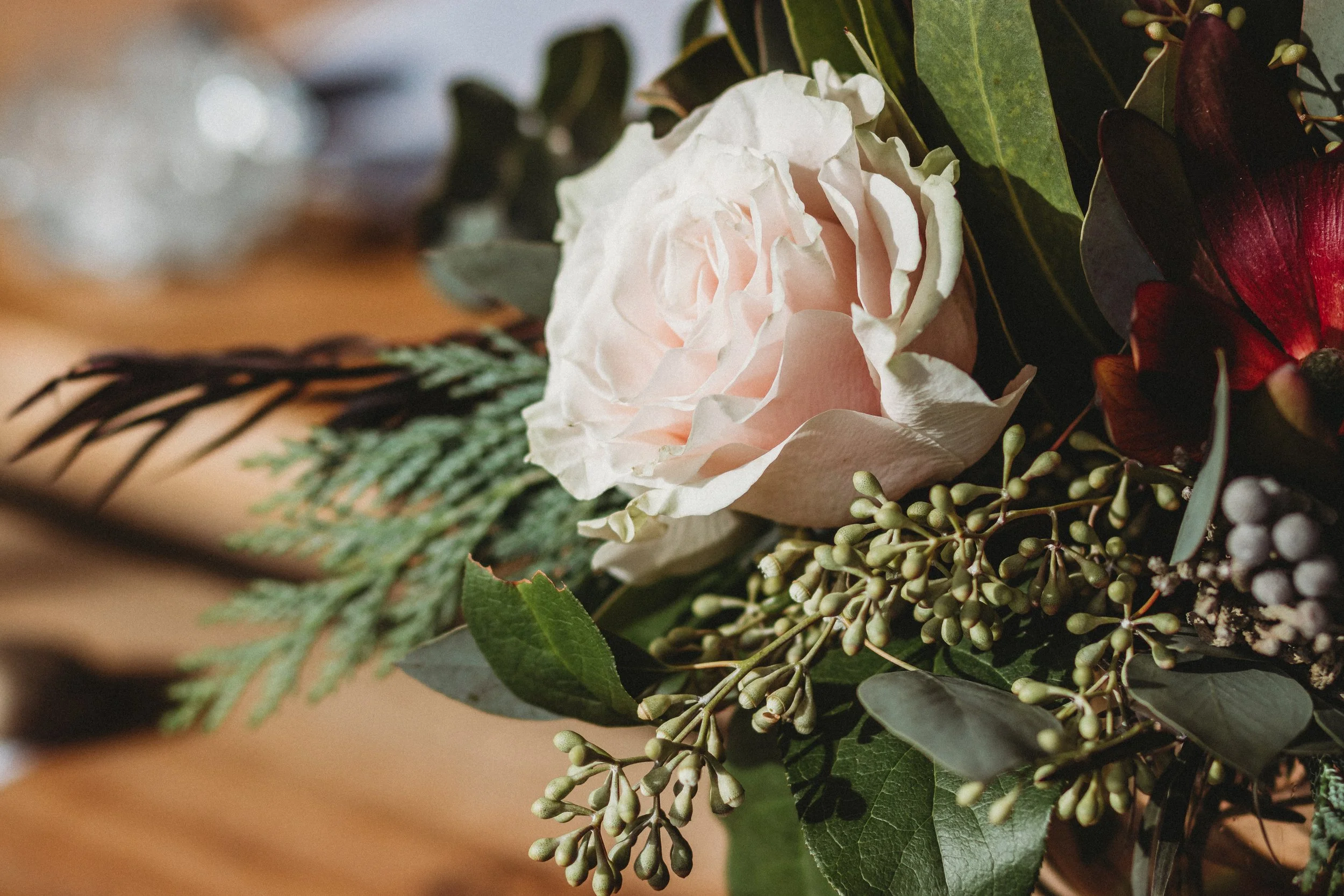 Close-up of a floral arrangement with a pink rose, green foliage, and small berry-like plants on a wooden surface.