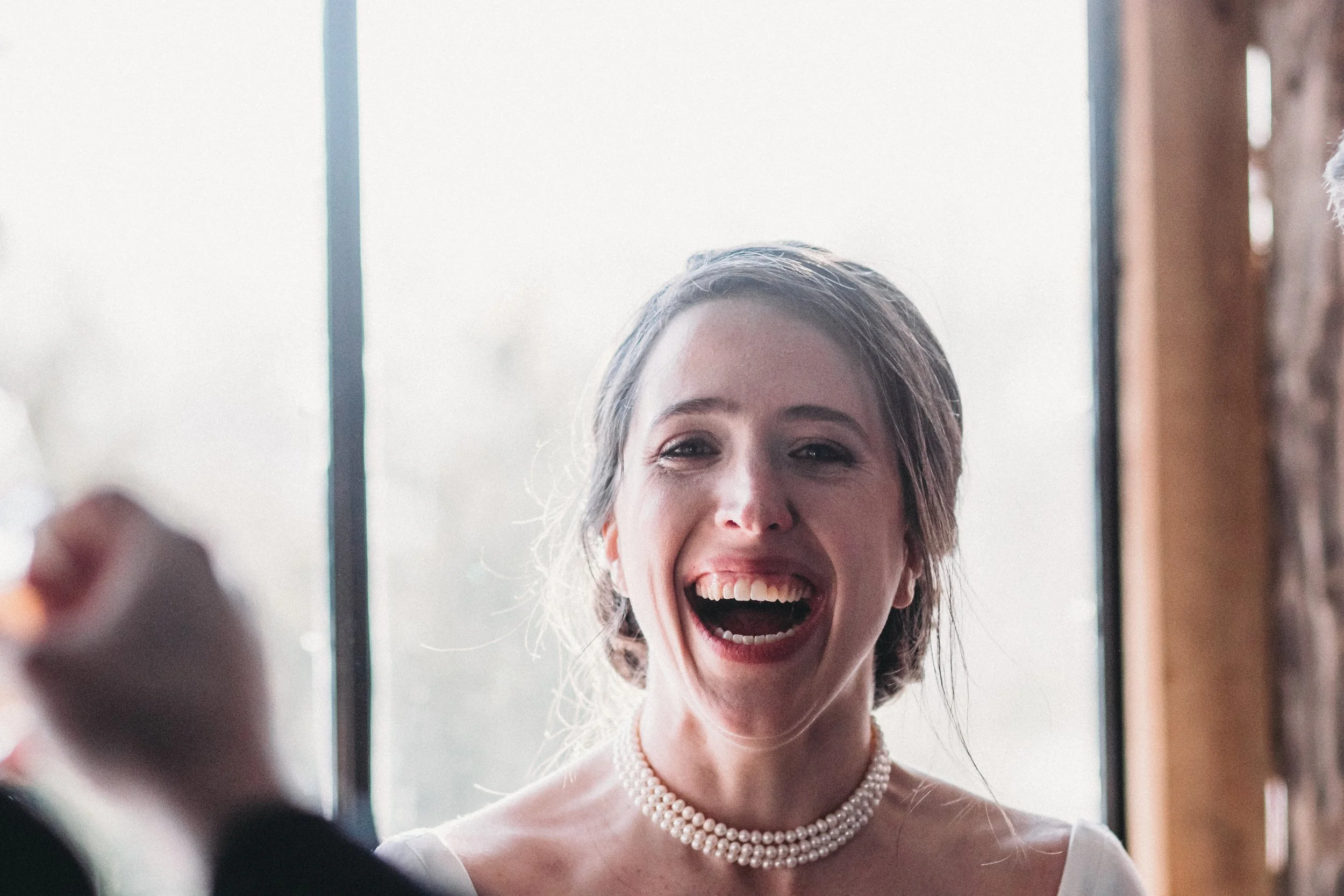 A woman with short brown hair smiling broadly, wearing a pearl necklace and a white top, standing in front of a large window with natural light.
