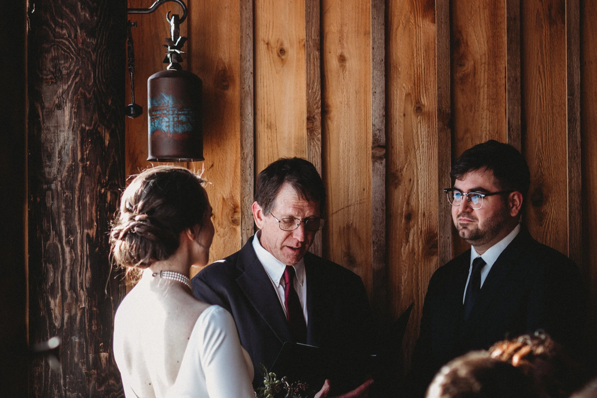 A wedding ceremony with a bride, groom, and officiant in front of a wooden wall, with a hanging bell or lantern above them.