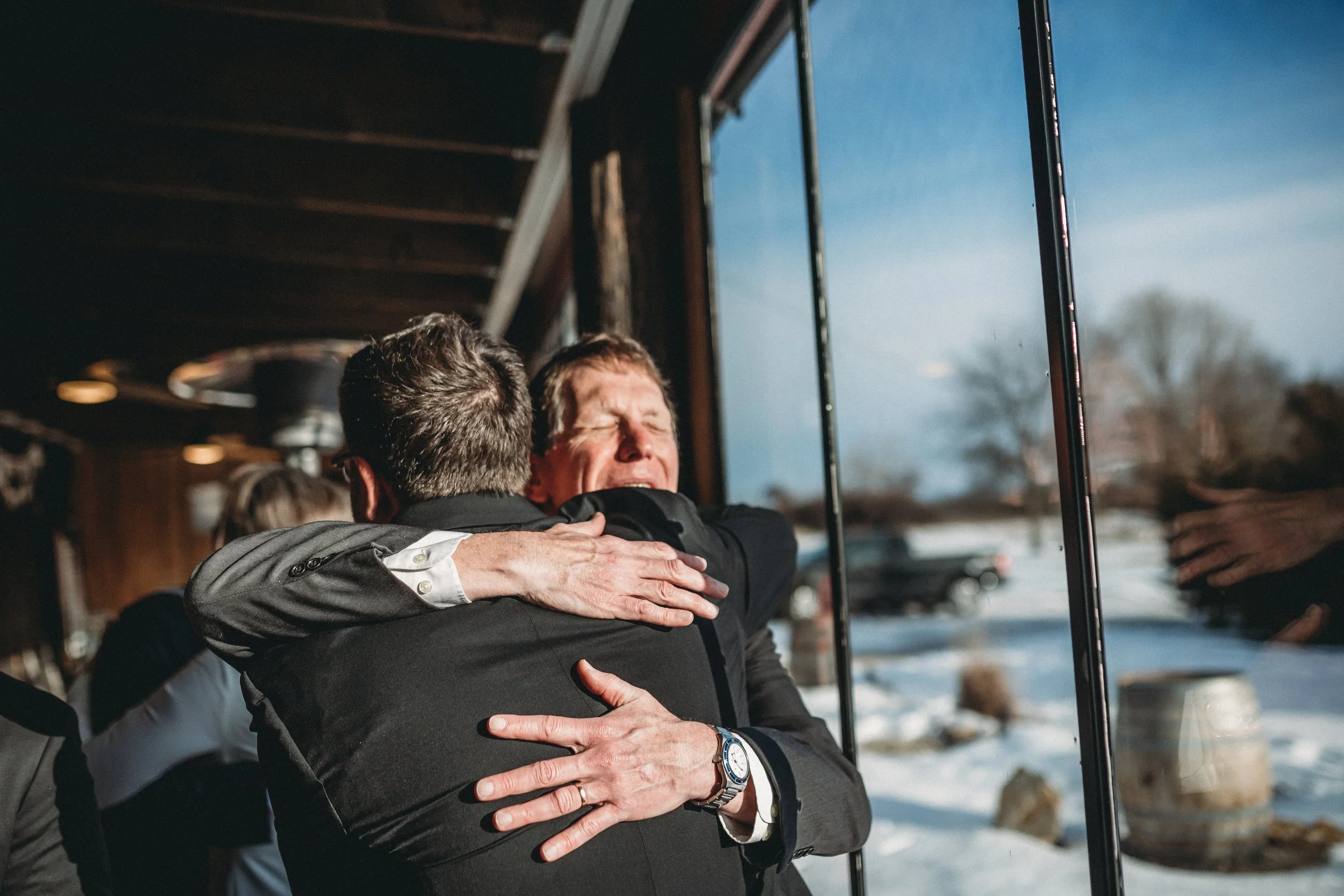 Two men hugging each other warmly in an indoor setting with large windows showing a snowy outdoor landscape and a clear blue sky.