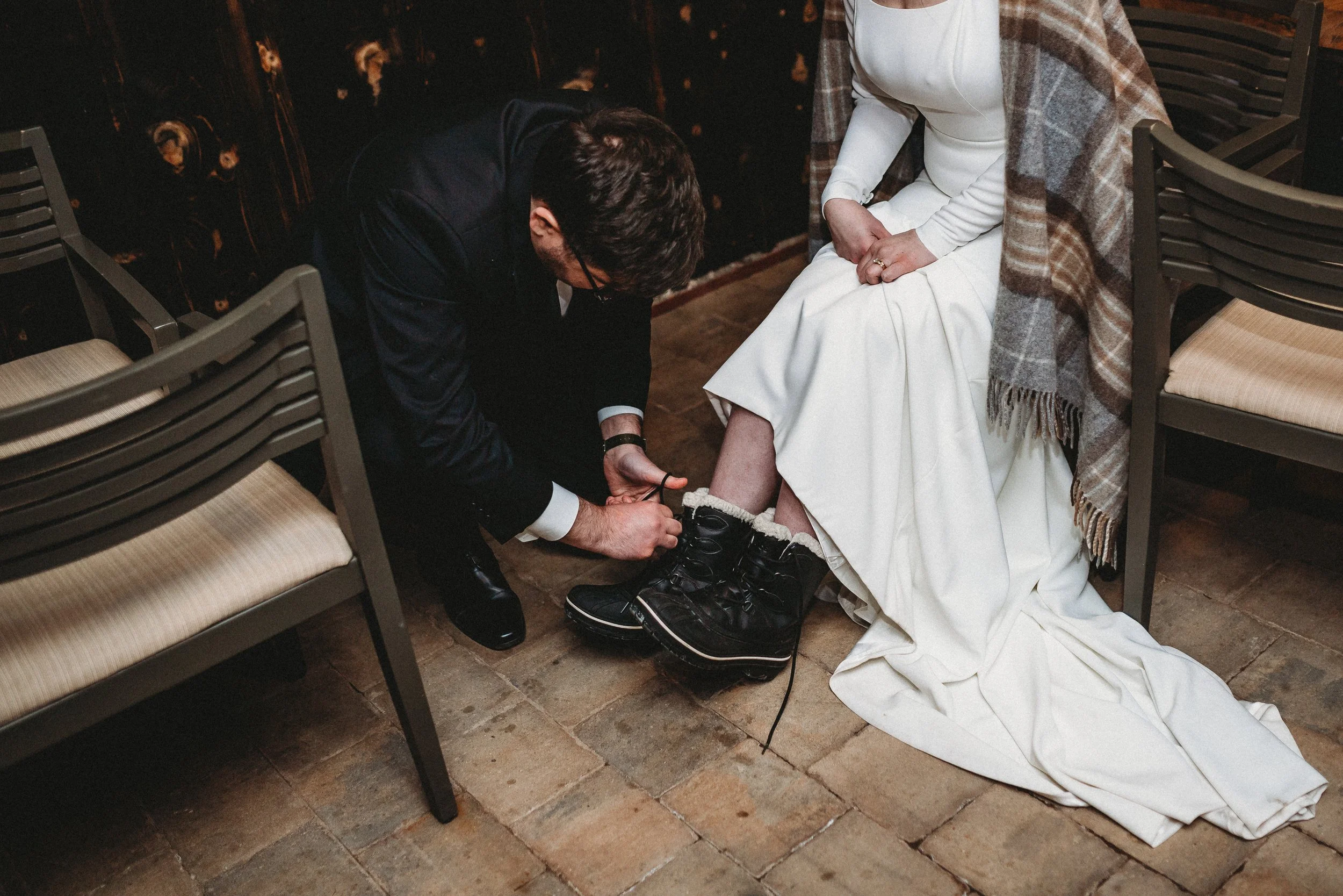 A man is helping a woman put on winter boots. The woman is sitting on a chair with a white dress and a plaid shawl, while the man kneels in front of her tying her shoe.