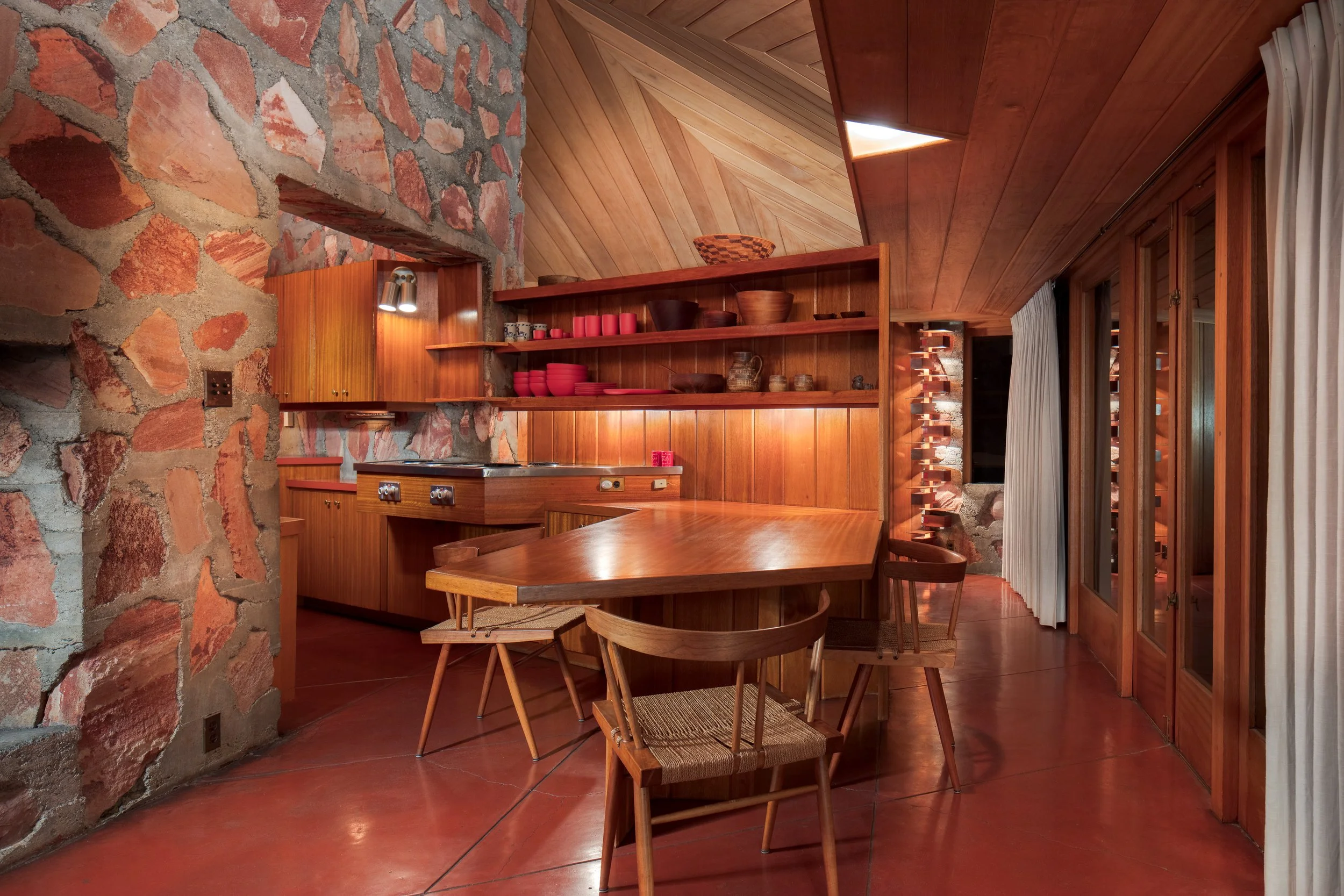 Midcentury modern dining room with a stone wall, wood-paneled ceiling, built-in shelves, dining table, and sculptural staircase