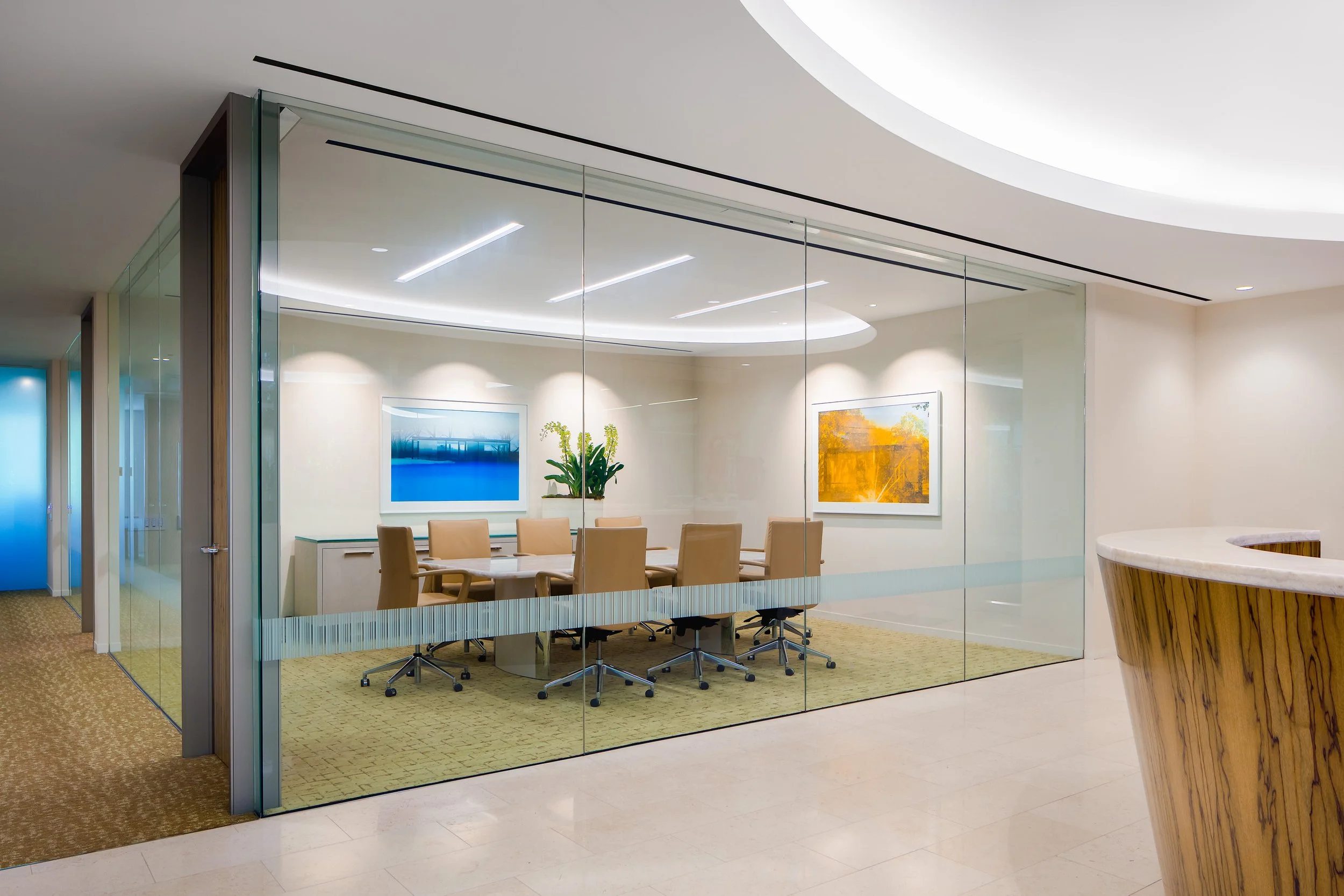 Modern glass-walled conference room with a long table, office chairs, artwork, and a plant beside a curved reception desk