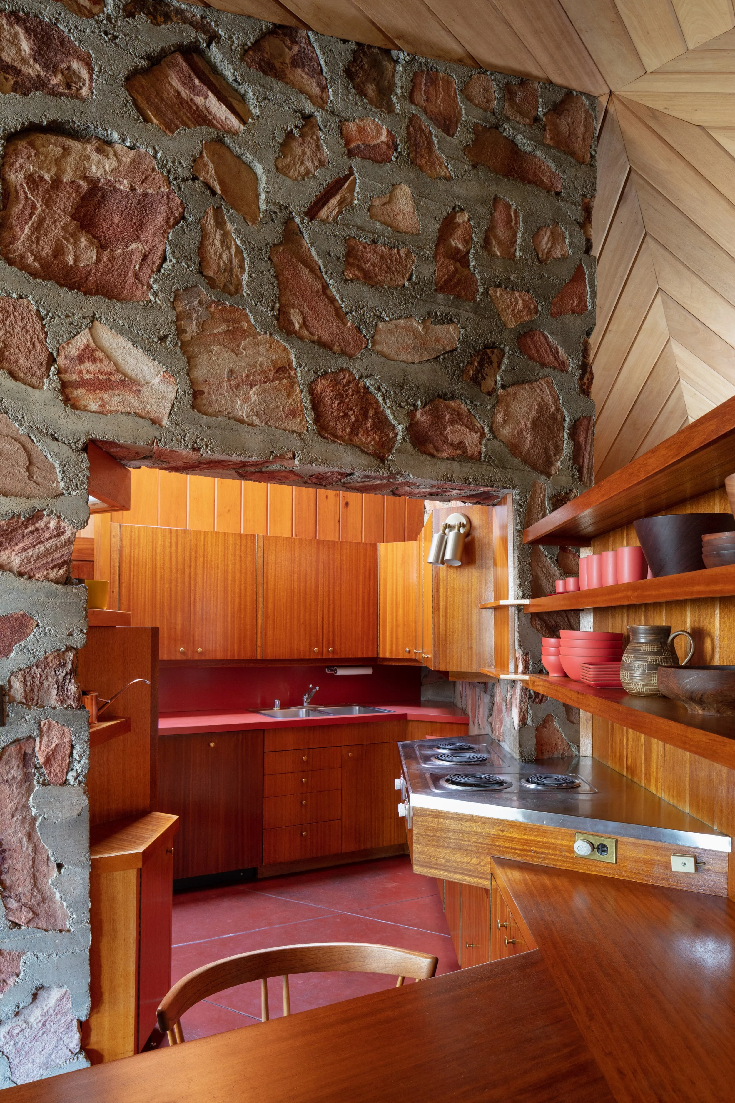 Rustic kitchen with exposed stone walls, wood cabinetry, red countertops, open shelves, and a freestanding electric stove.