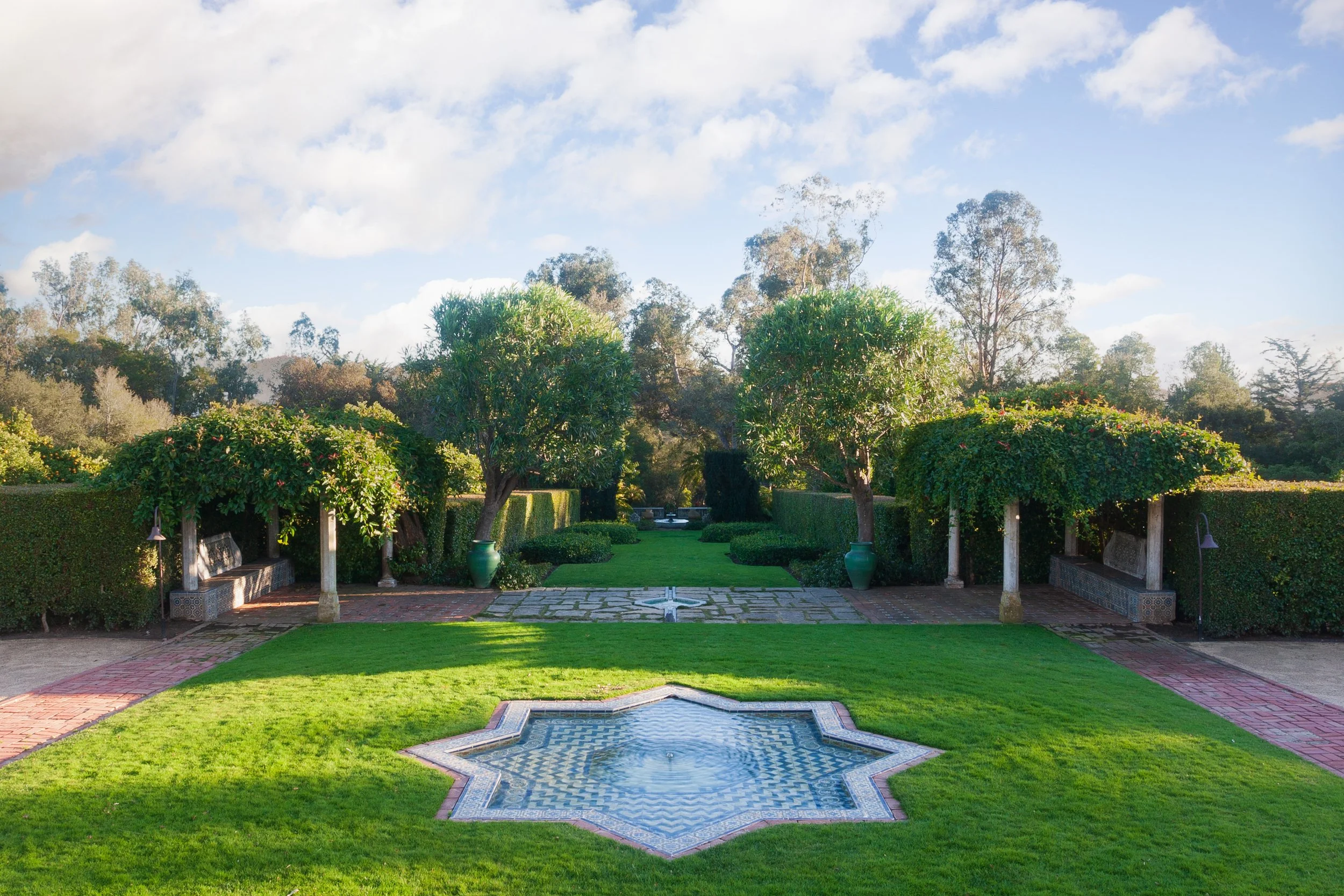 Formal landscaped garden with manicured hedges, topiary trees, tiled fountain, brick paths, and benches under a sunny sky
