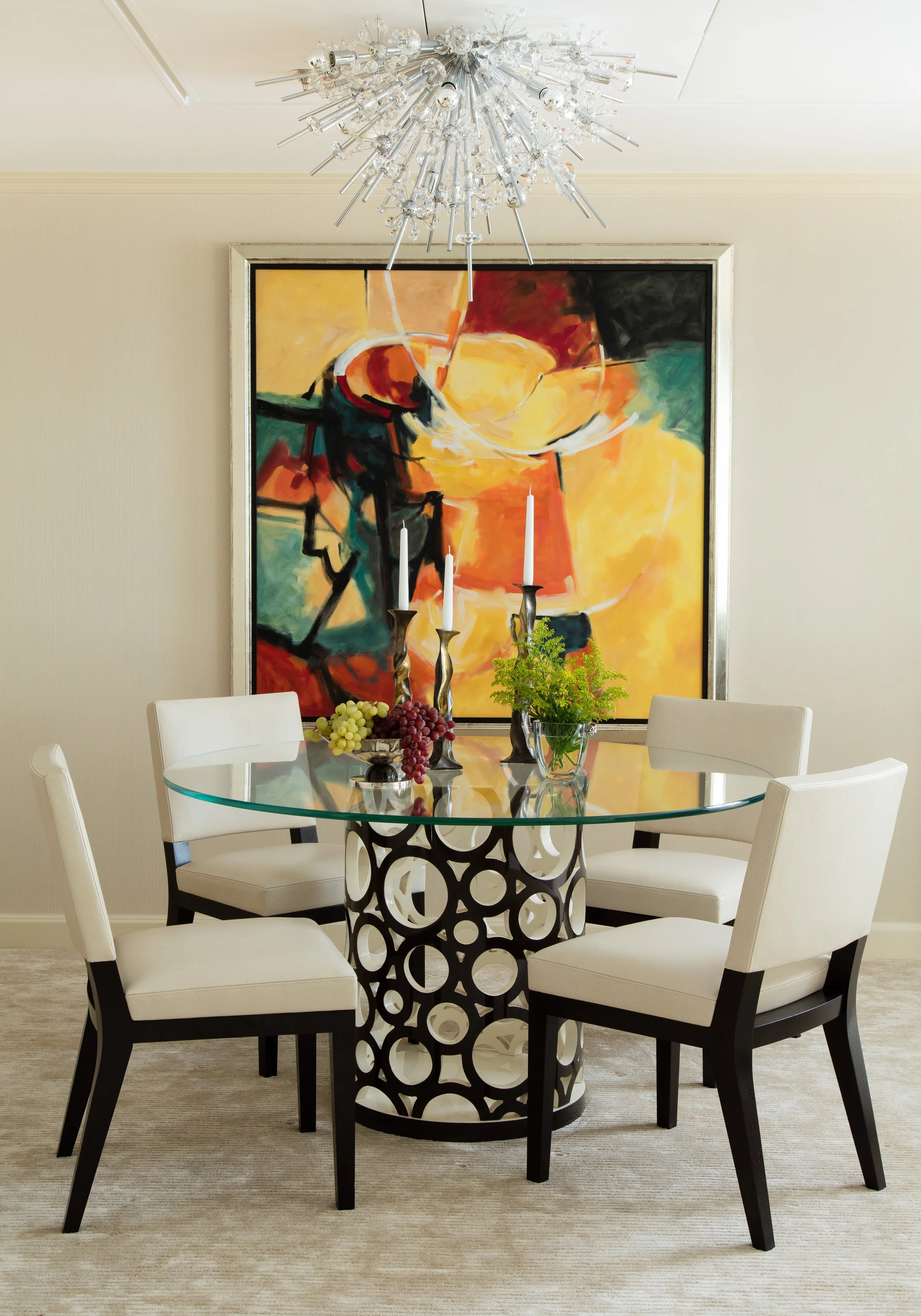 Elegant dining room with a glass table, white chairs, colorful abstract painting, crystal chandelier, candles, grapes, and floral centerpiece