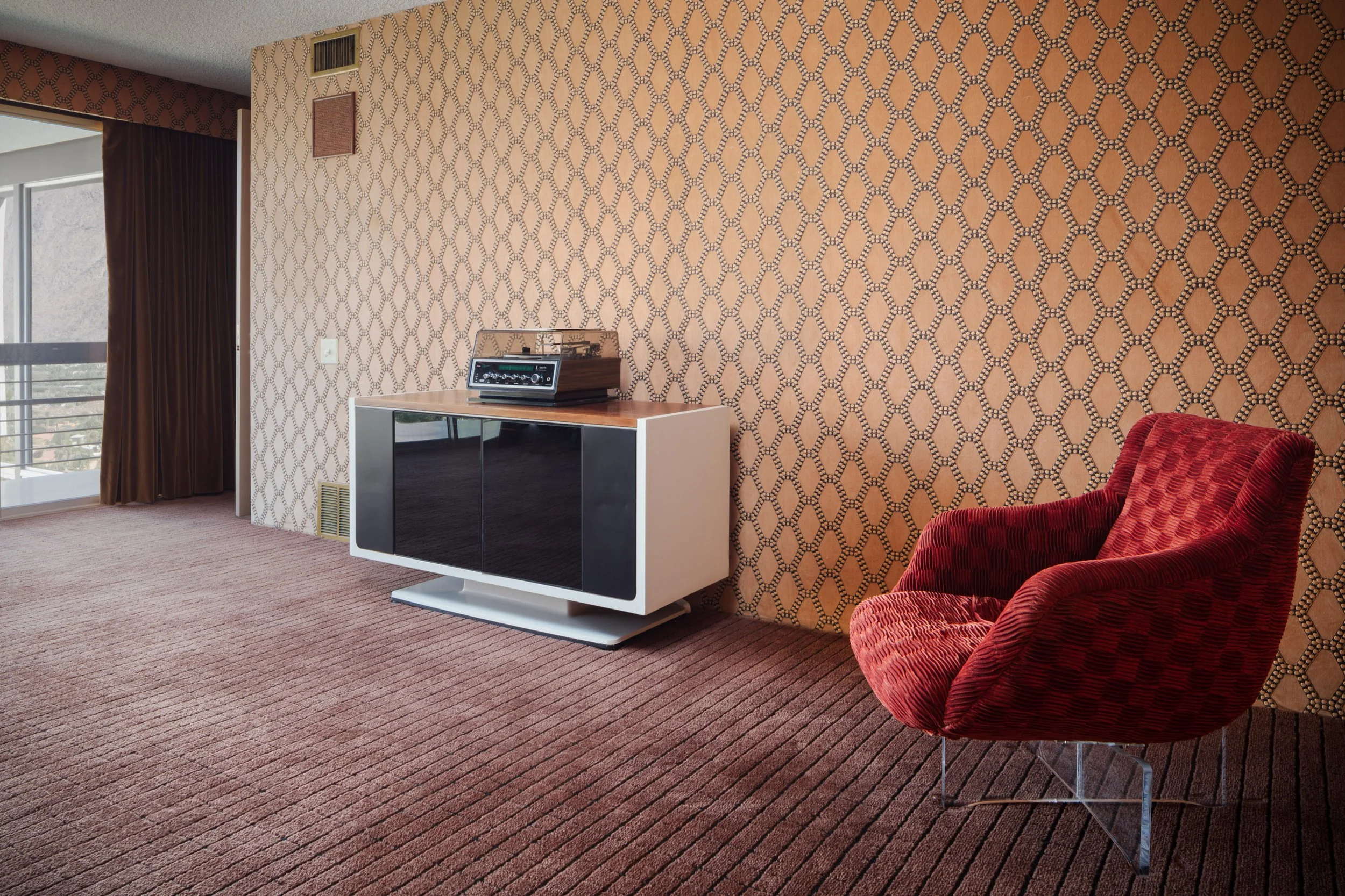 Mid-century modern living room with geometric patterned wallpaper, vintage stereo console, red lounge chair, and floor-to-ceiling windows.