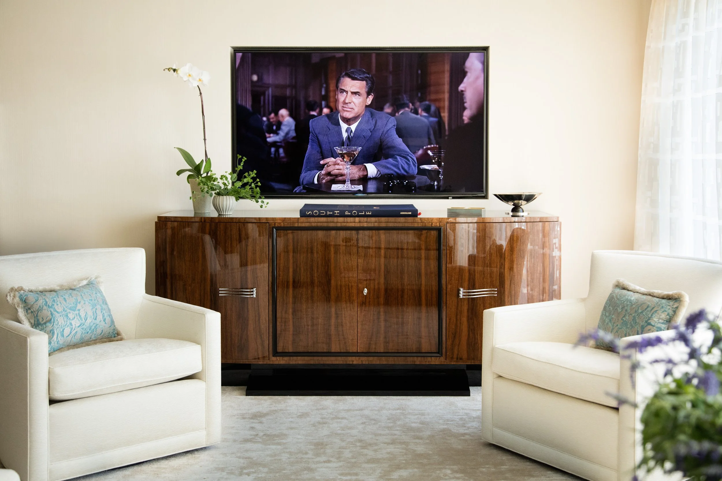 Elegant living room with a wall-mounted TV showing Cary Grant, white armchairs, a wood credenza, orchid, and decorative bowl