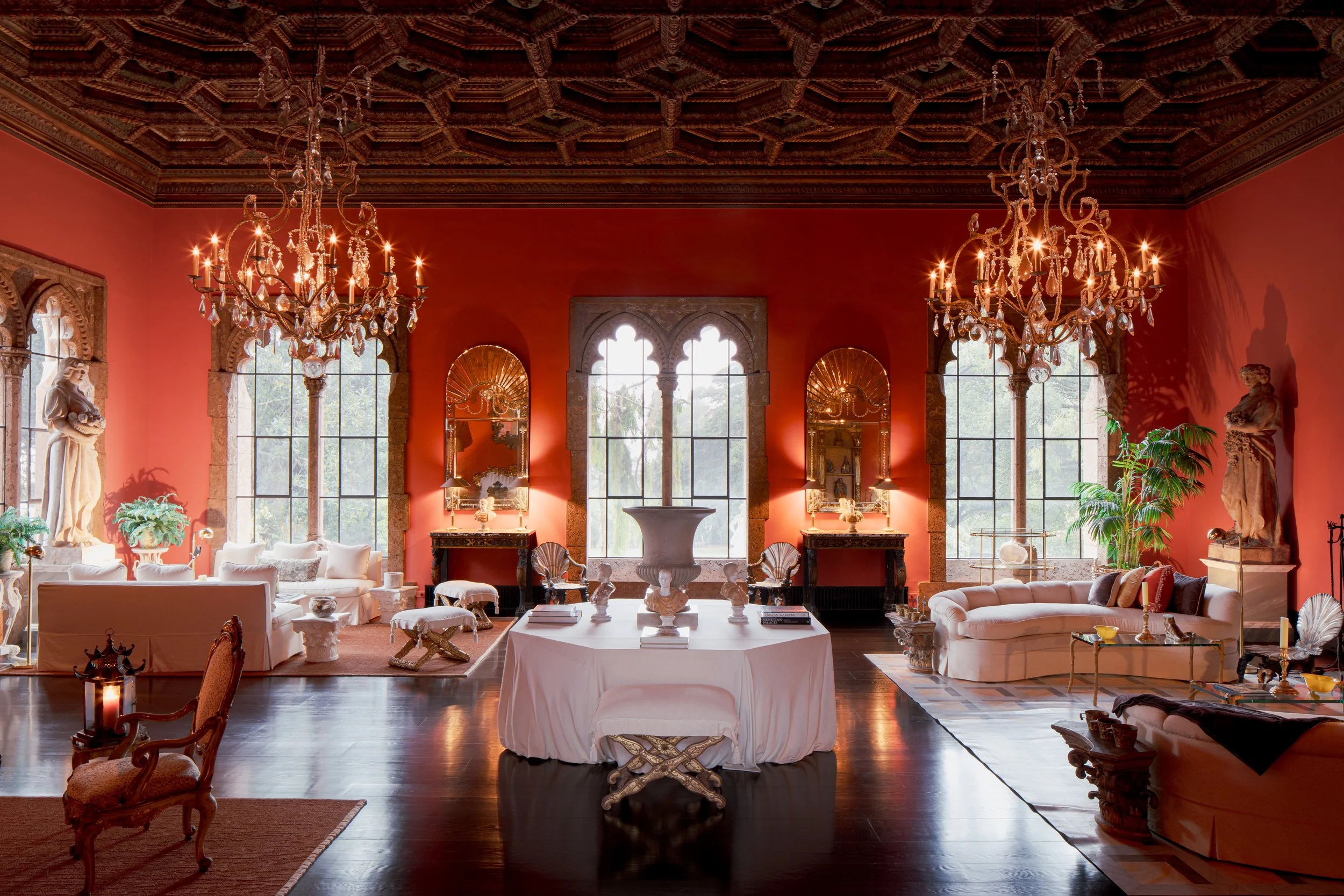 Grand red luxury living room with ornate carved wood ceiling, crystal chandeliers, arched windows, antiques, sculptures, and white sofas