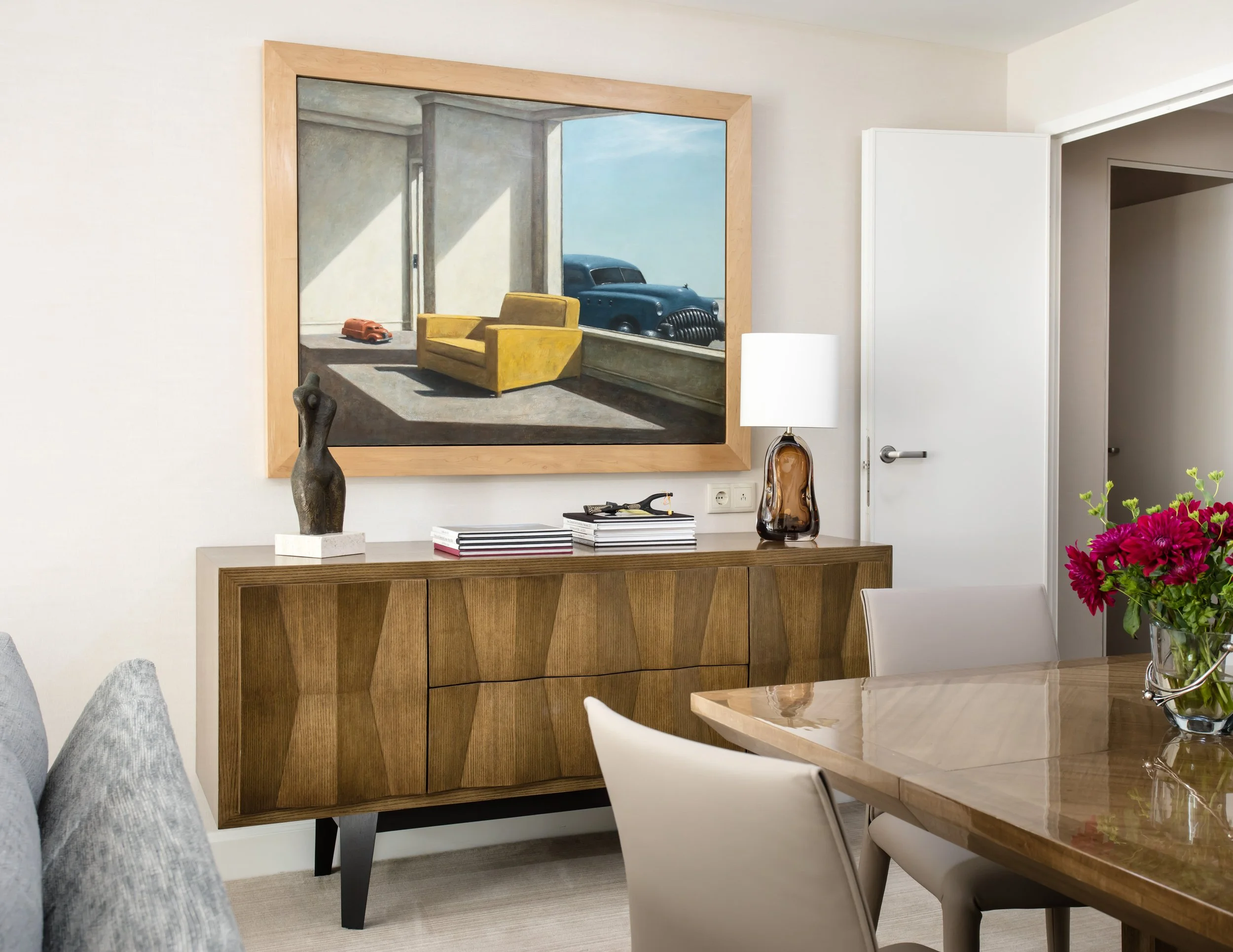 Modern dining room with a wood sideboard, framed vintage car artwork, sculptural decor, lamp, and vase of flowers