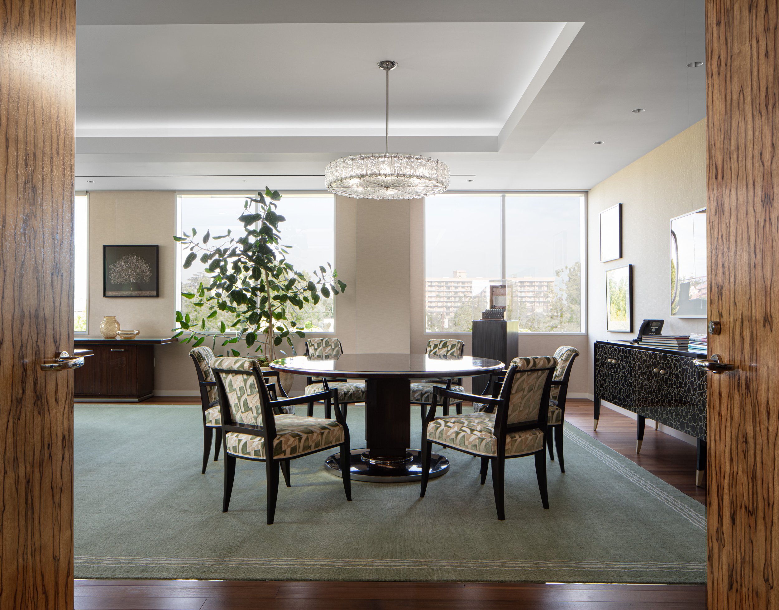 Elegant modern dining room with a round table, upholstered chairs, crystal chandelier, large windows, and indoor tree