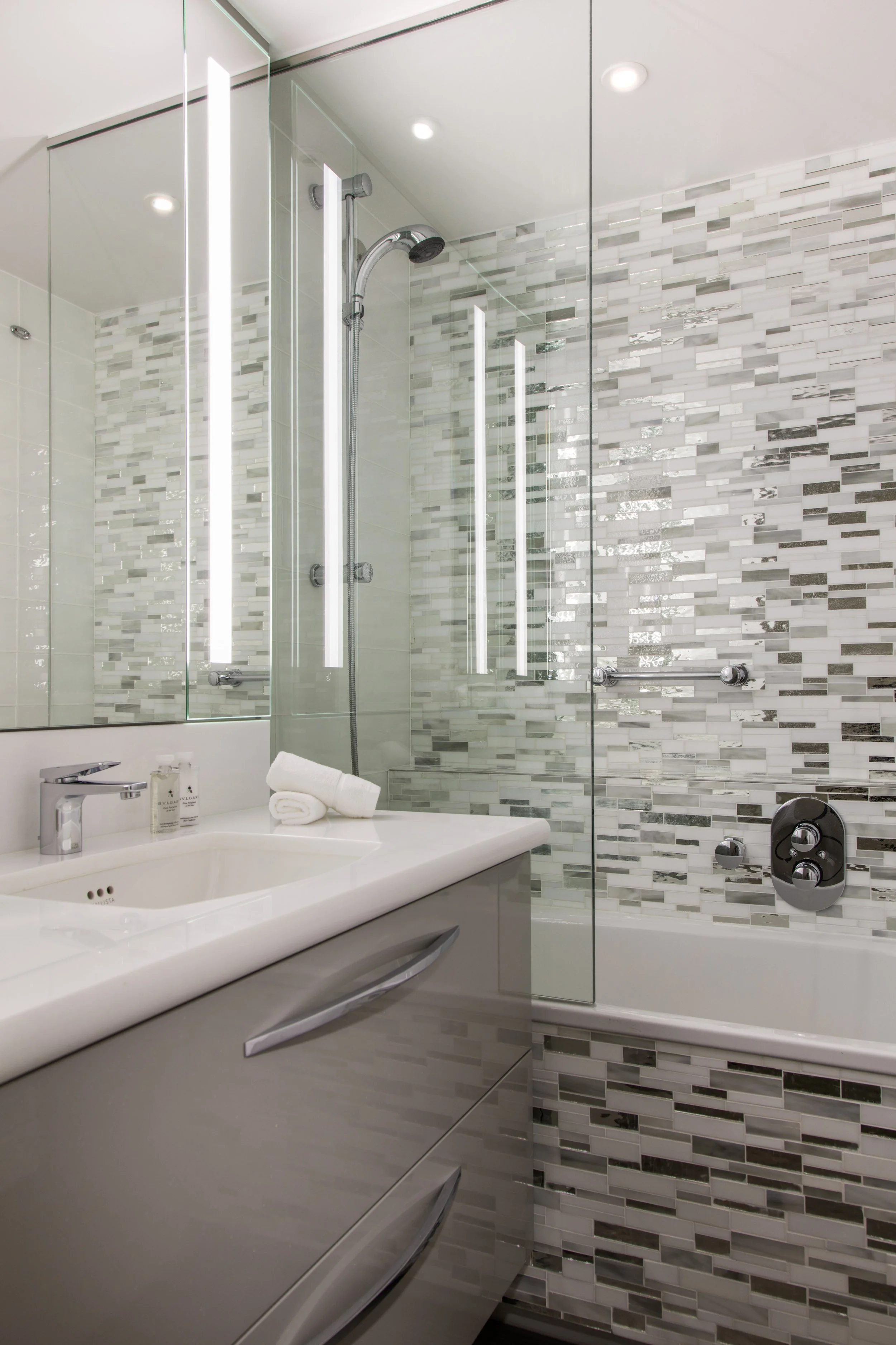 Modern bathroom with a glass-enclosed shower, freestanding bathtub, chrome fixtures, mosaic tile walls, and rolled towels