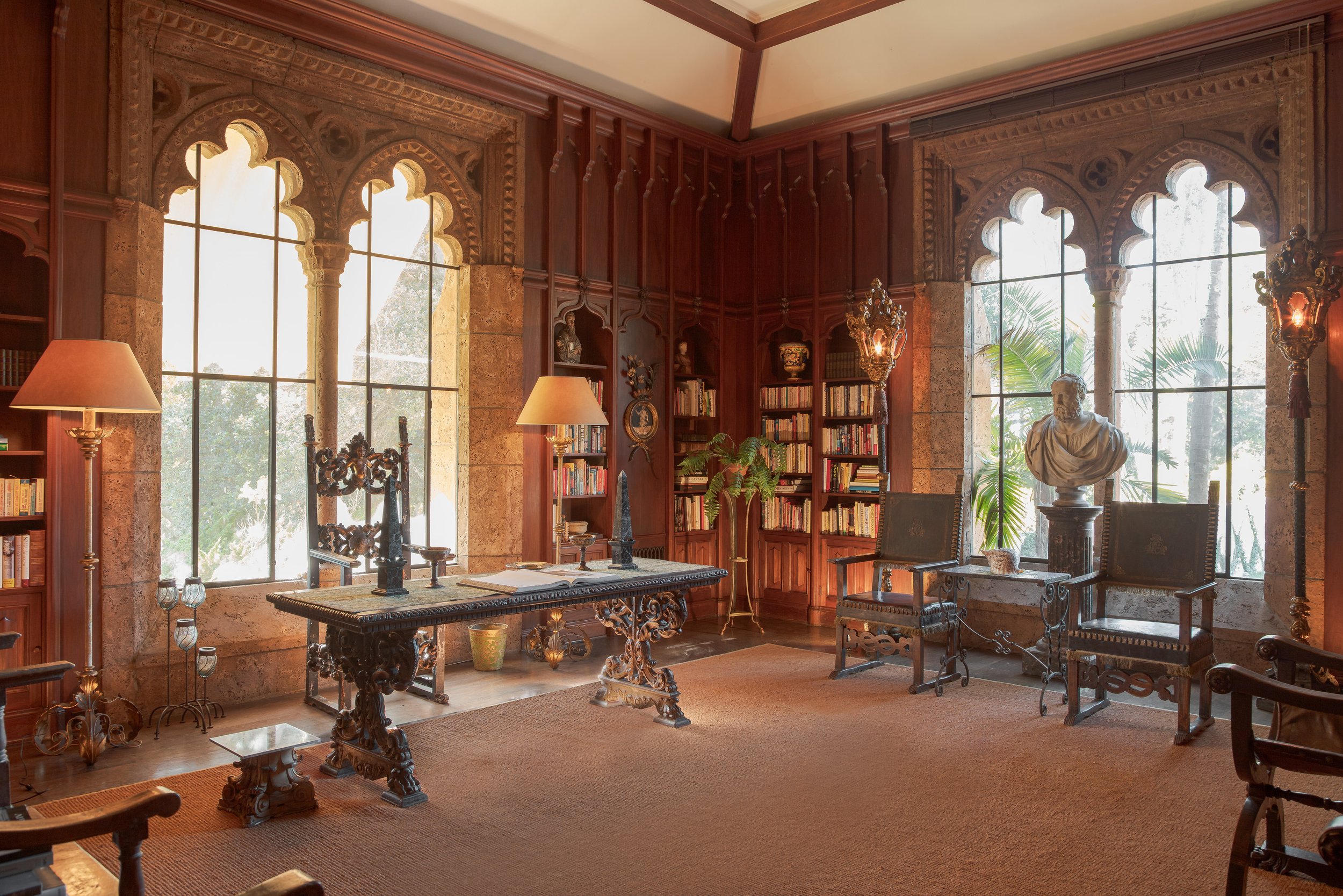 Elegant wood-paneled library with carved stone Gothic windows, bookshelves, ornate table, lamps, and classical bust sculpture