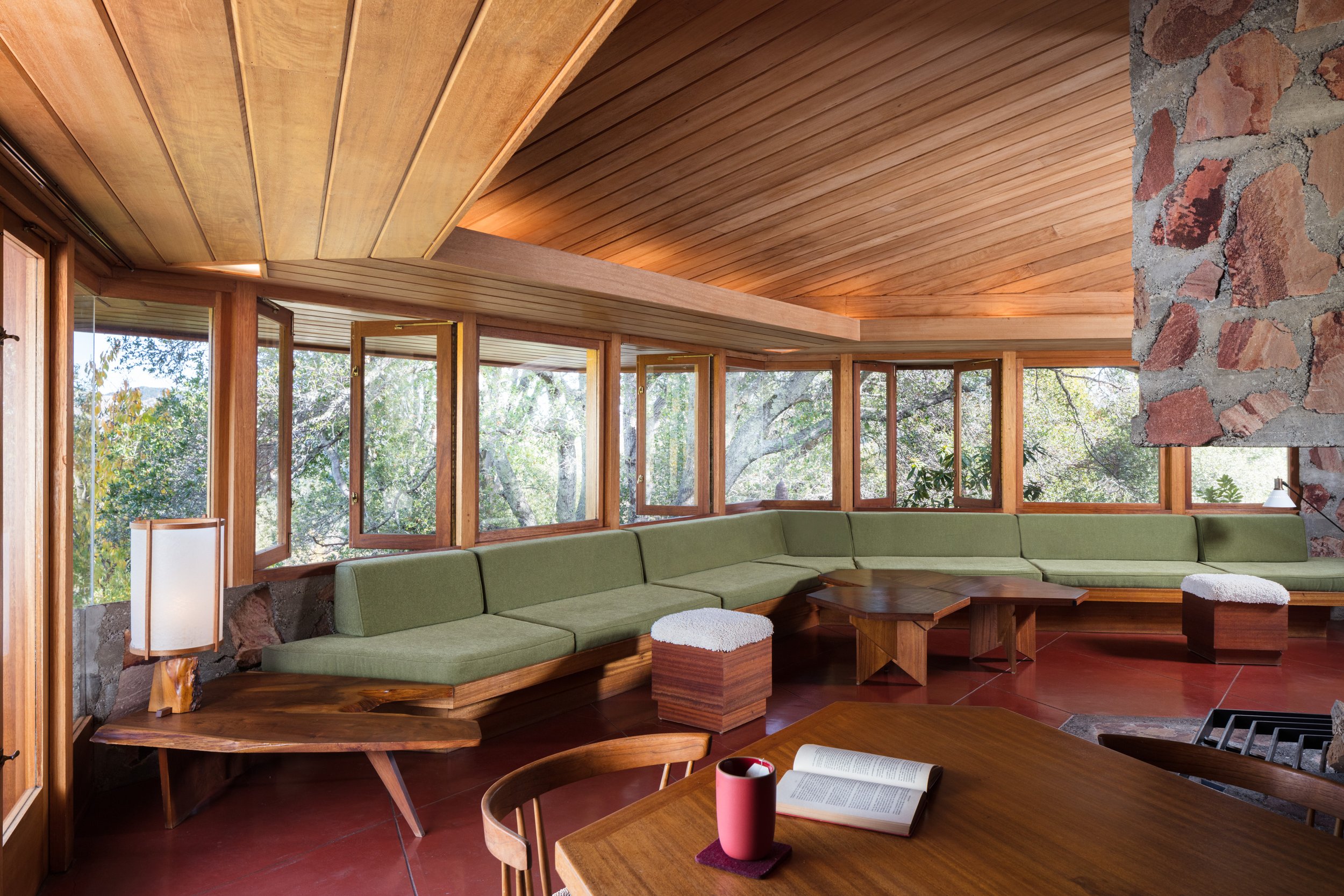 Midcentury modern living room with wood-paneled ceiling, floor-to-ceiling windows, green sectional sofa, stone fireplace, and wooden coffee tables