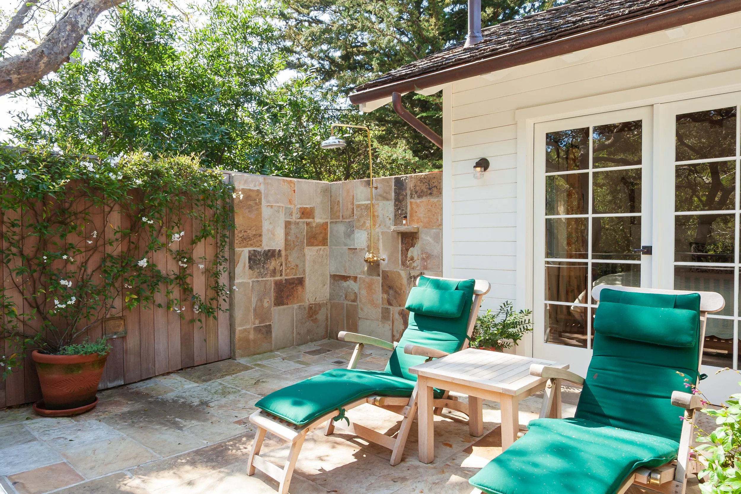 Sunny private patio with teal lounge chairs, a wooden table, stone outdoor shower, flowering vines, and glass French doors.
