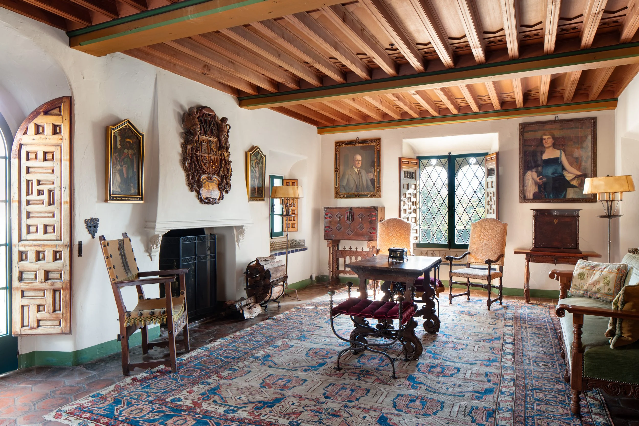 Historic Spanish-style living room with exposed wood-beam ceiling, fireplace, antique furniture, portraits, and patterned Persian rugs