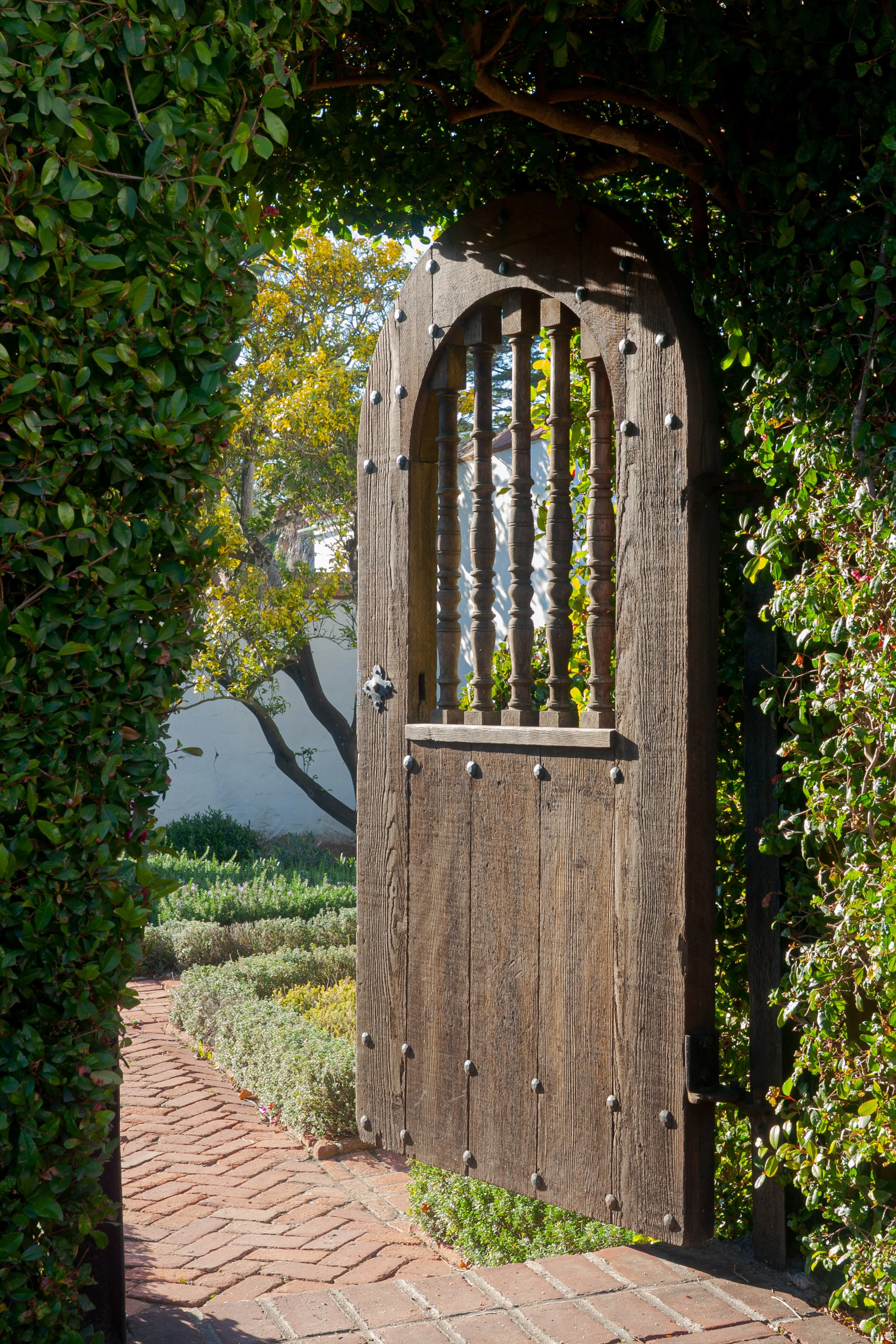 Rustic wooden garden gate with decorative metal studs, surrounded by lush greenery and opening onto a brick walkway