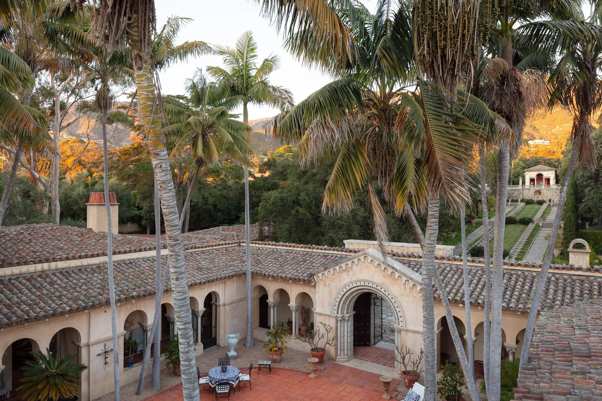 Spanish-style villa courtyard with terracotta tile roofs, tall palm trees, arched walkways, landscaped gardens, and sunlit mountains in the background