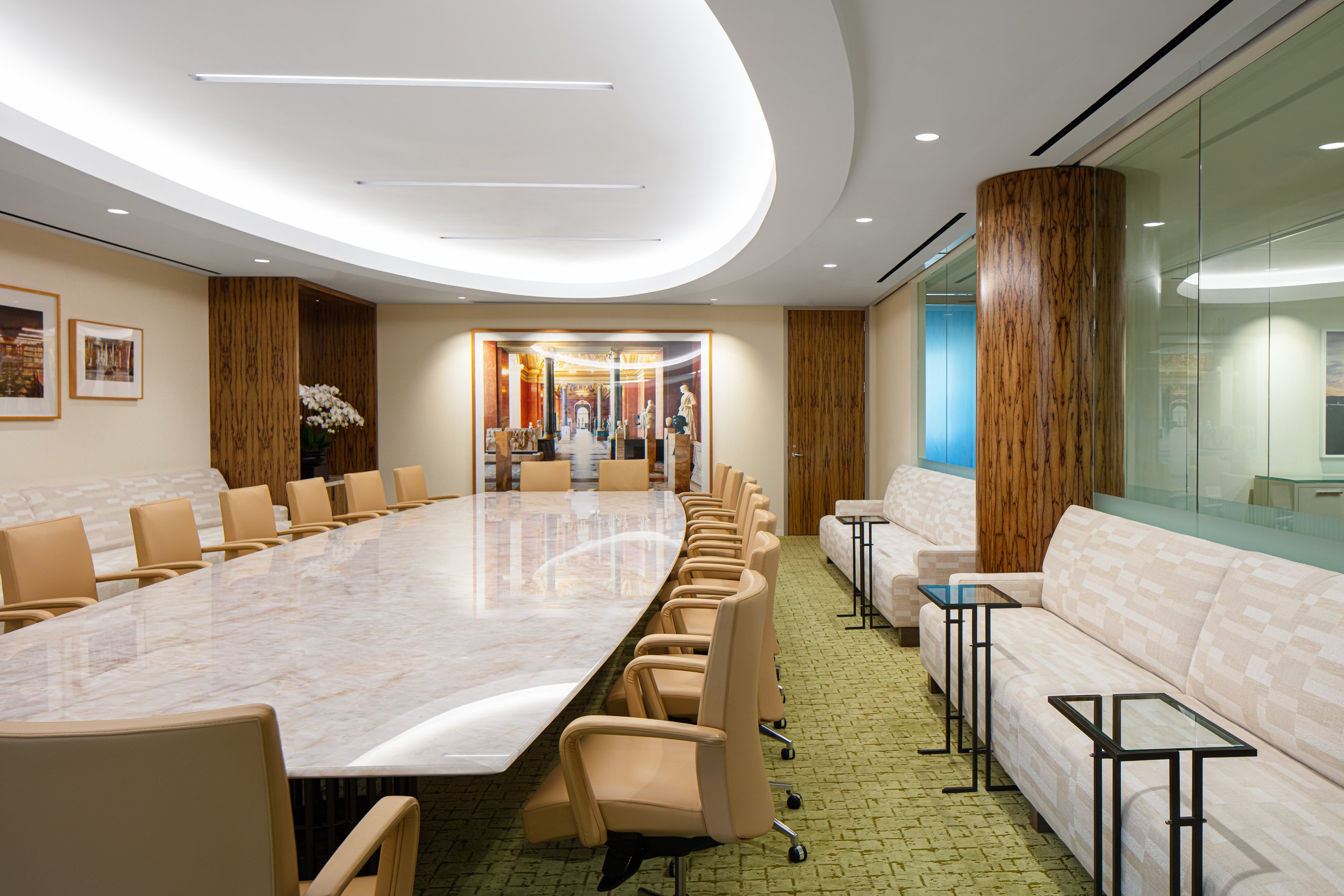 Modern executive conference room with a large marble table, tan chairs, wood-paneled columns, sofas, and glass walls