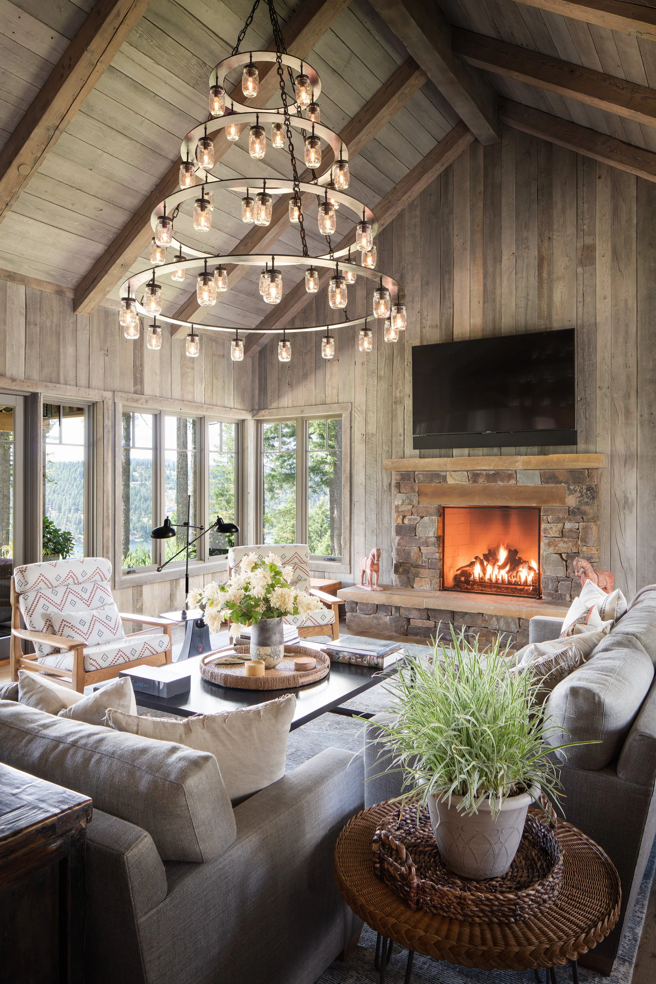 Rustic mountain lodge living room with a stone fireplace, glowing fire, mason jar chandelier, wood-paneled walls, and large windows overlooking a forested landscape.