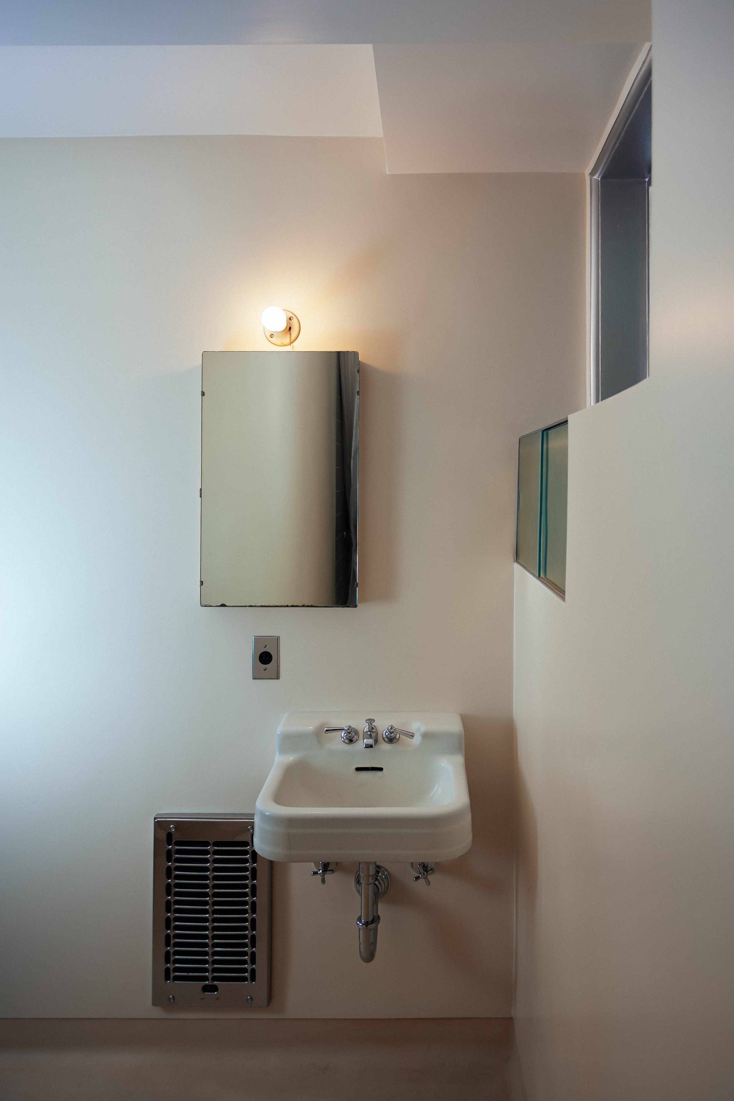 Close-up of a vintage bathroom with a wall-mounted sink, mirror, light fixture, and ventilation grille