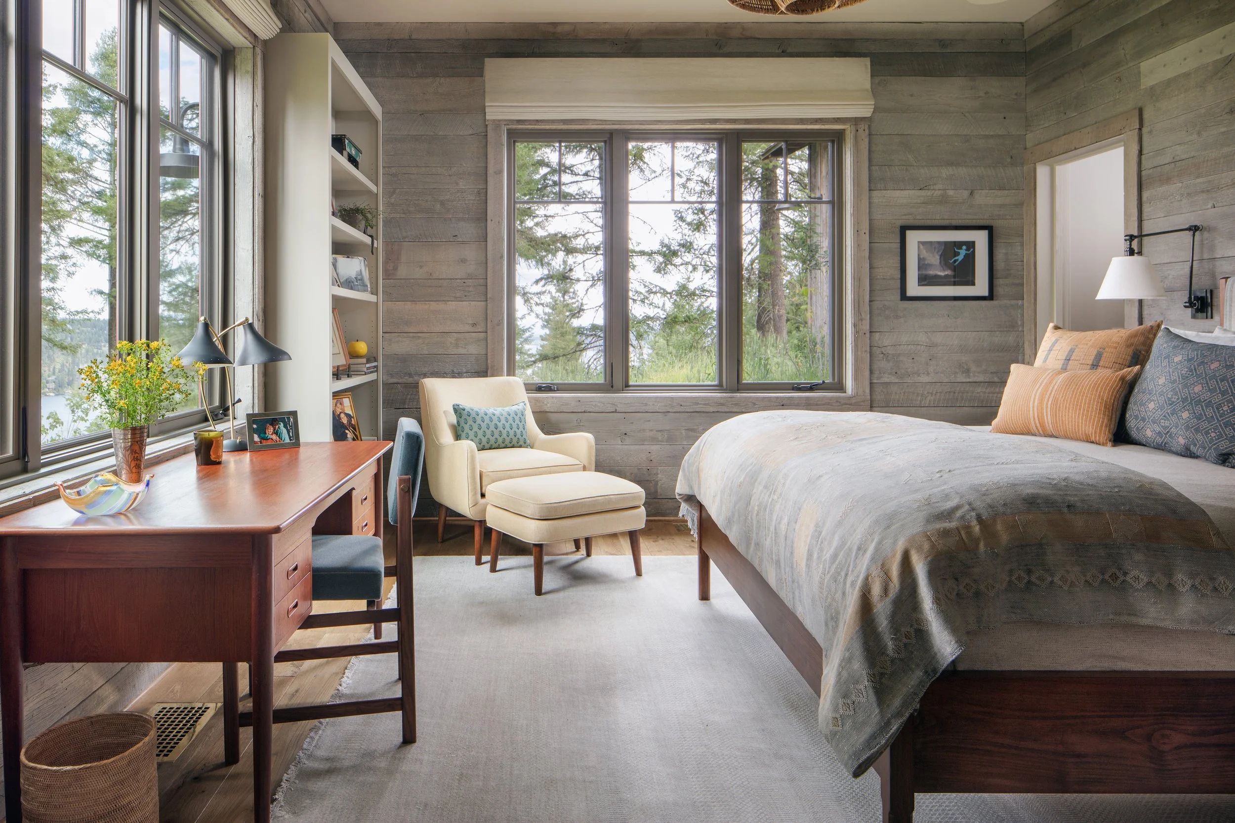 Rustic modern bedroom with wood-paneled walls, large windows overlooking a forested lake, a bed, armchair, desk, and shelving.