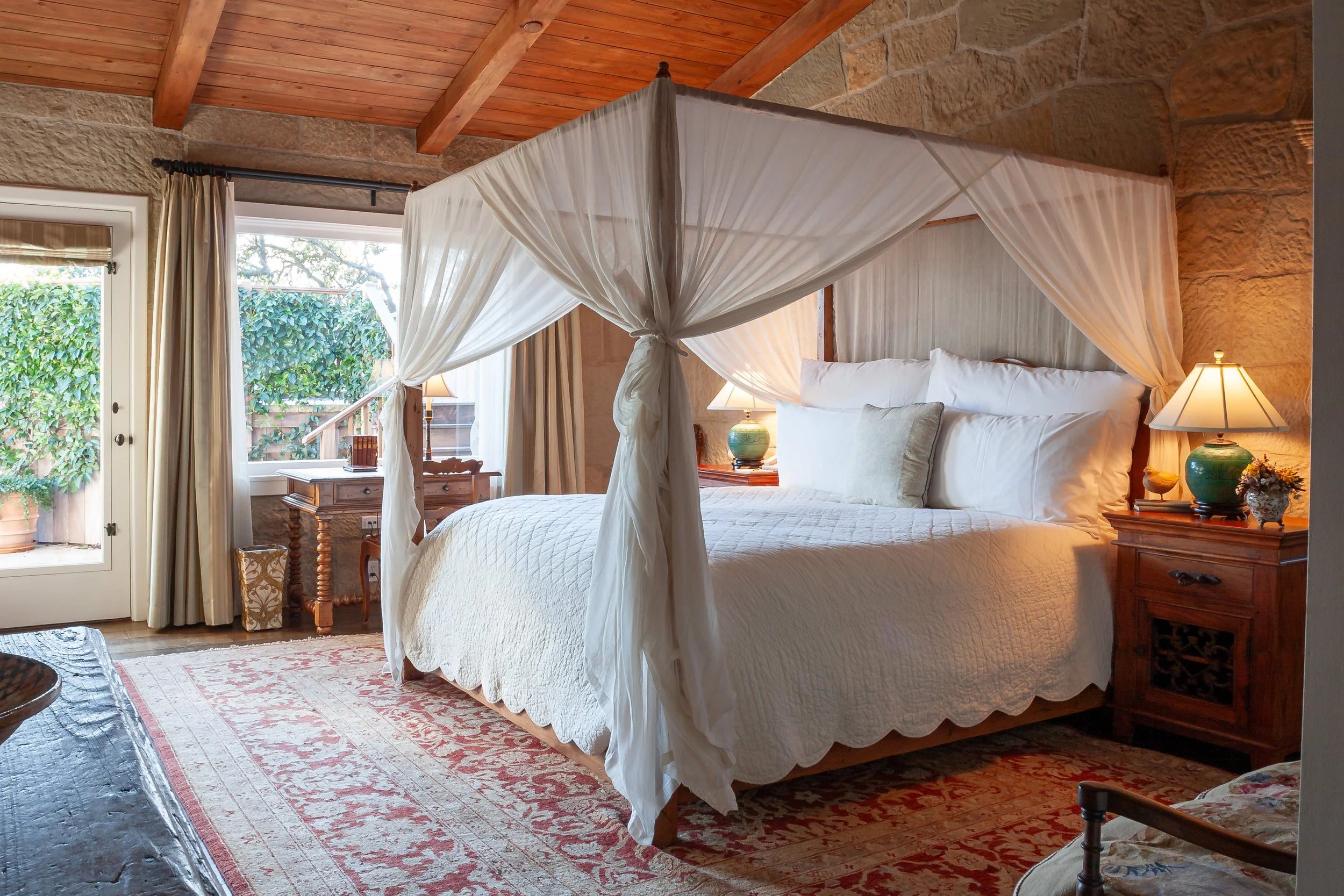 Rustic stone bedroom with a white canopy bed, wooden ceiling beams, bedside lamps, and patterned area rug