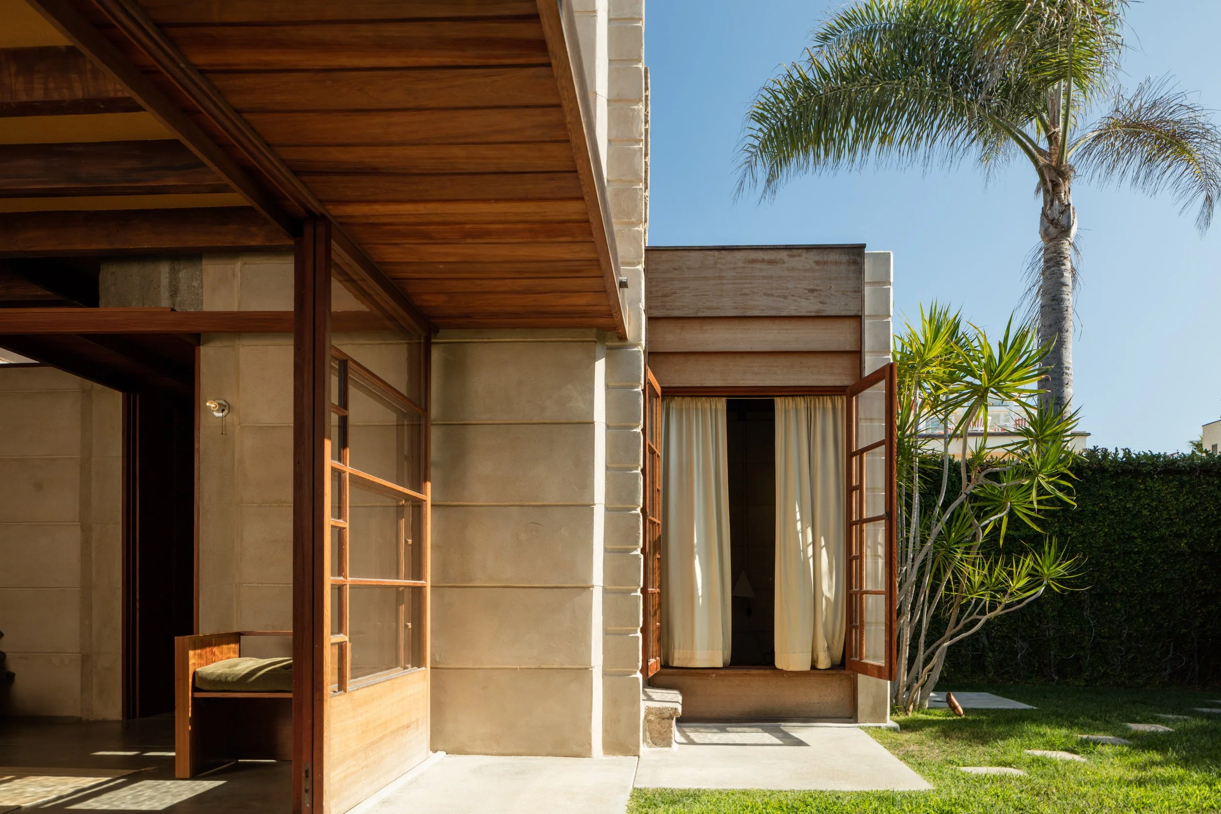 Modern stucco house with open wooden doors, a covered patio, tropical palm trees, and a sunny lawn
