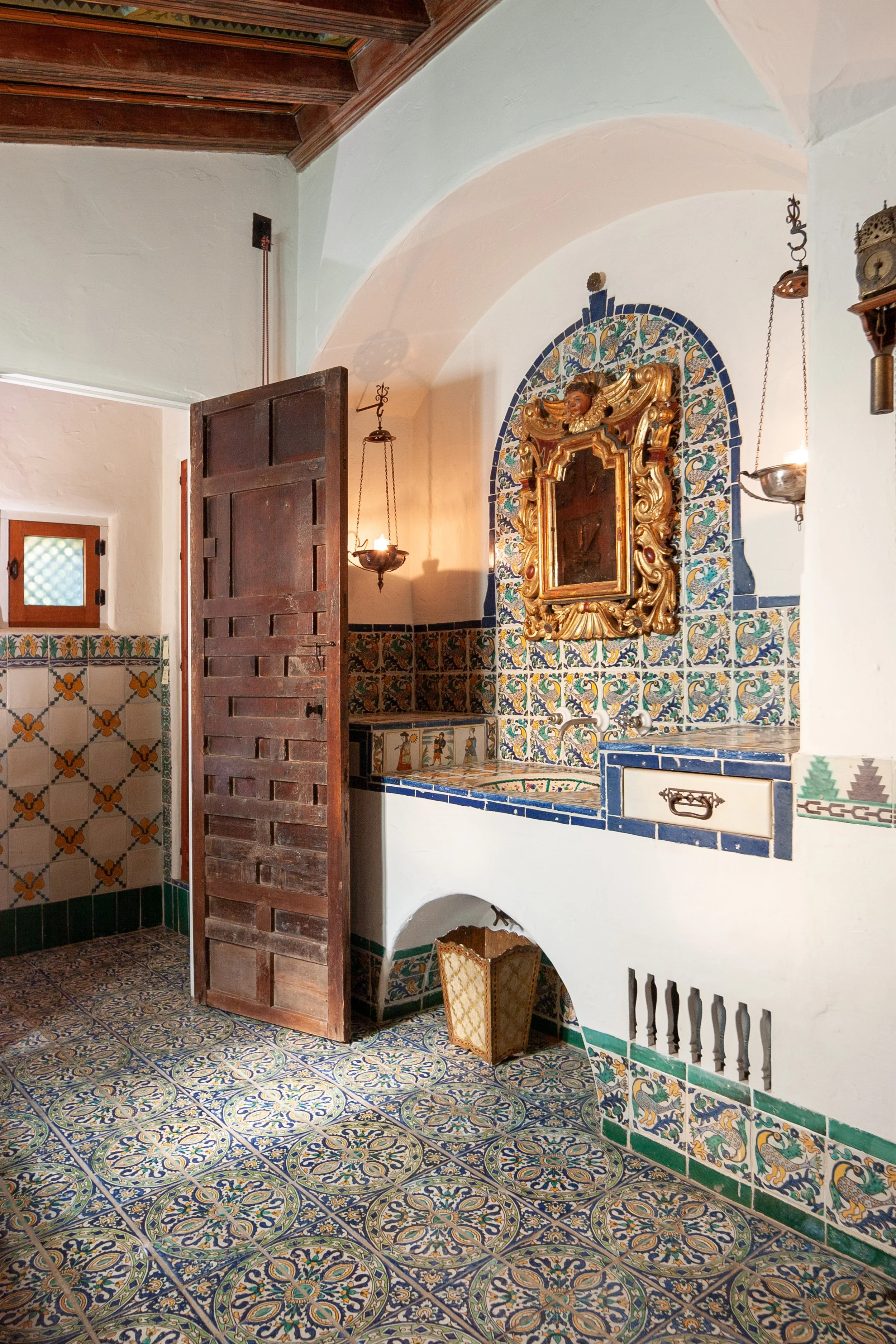 Ornate Spanish-style tiled interior with patterned ceramic floor and walls, a carved wooden door, arched alcove, gilded mirror, and hanging lanterns.