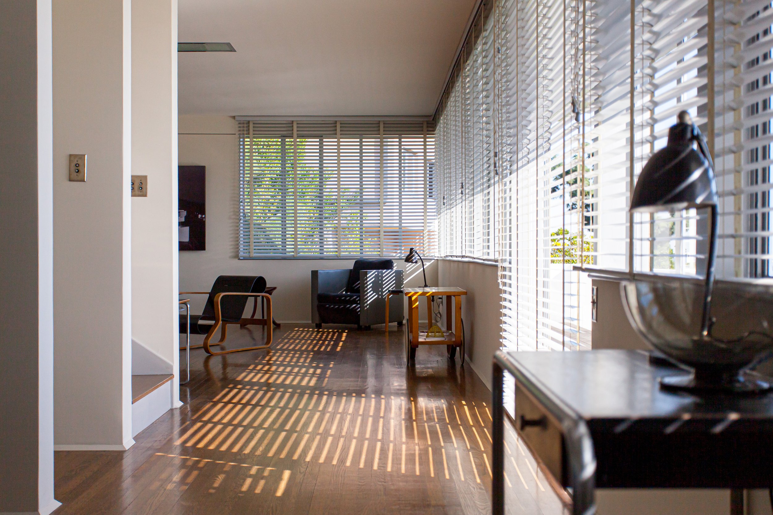 Sunlit modern living room with wood floors, lounge chairs, tables, and expansive windows covered by Venetian blinds.