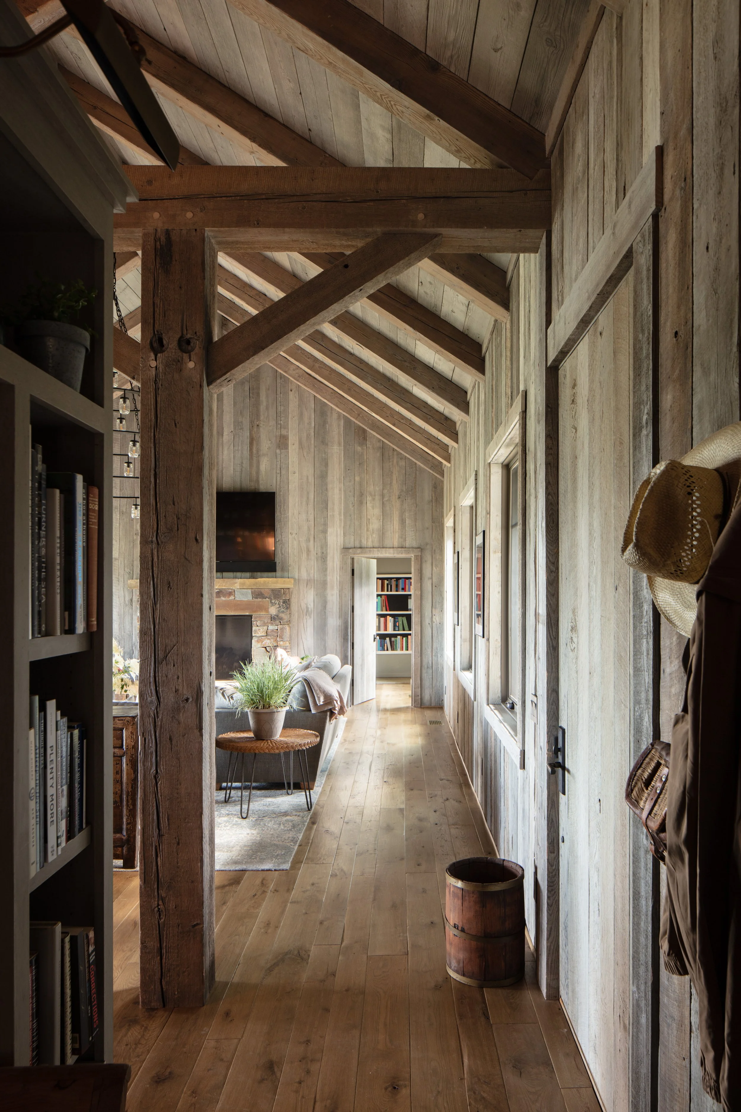 Rustic barn-style home interior with exposed timber beams, reclaimed wood walls, light wood floors, bookshelves, and a cozy living room.