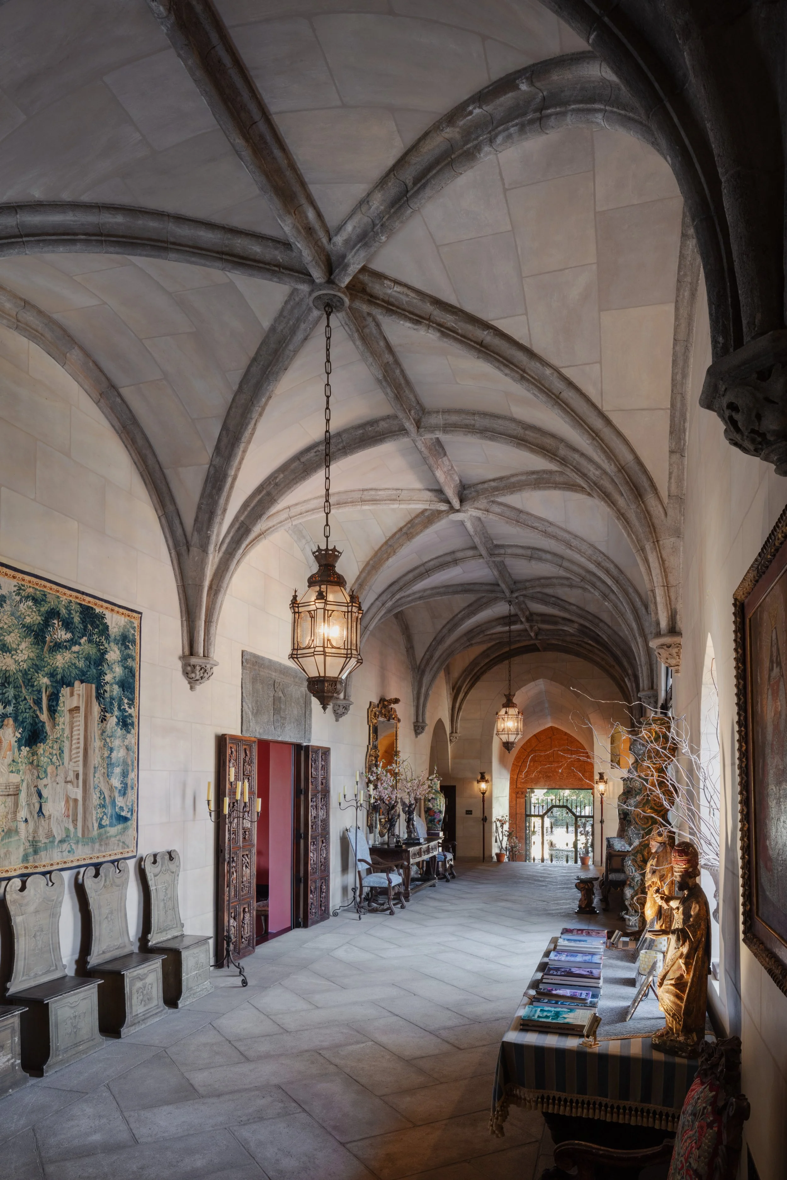 Ornate historic stone hall with a vaulted ribbed ceiling, hanging lanterns, tapestries, carved furniture, and religious artwork.