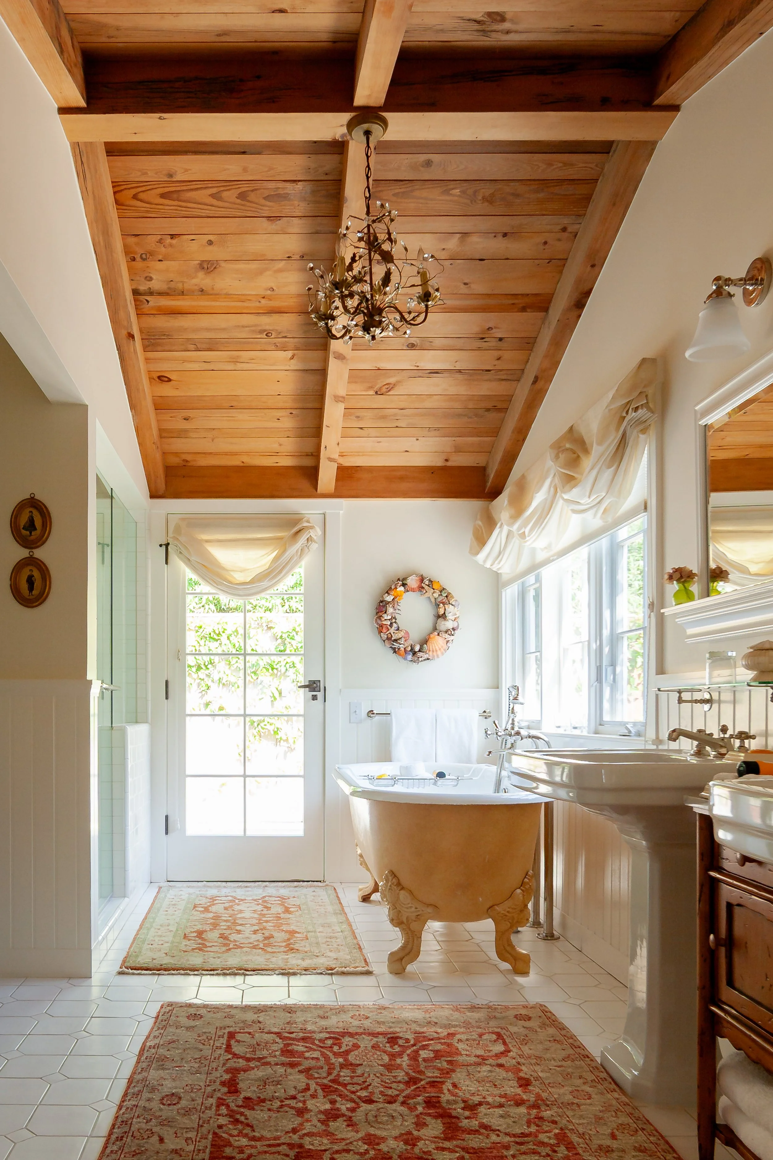 Elegant bathroom with exposed wood-beam ceiling, crystal chandelier, clawfoot bathtub, glass shower, patterned Persian rugs, and bright windows.