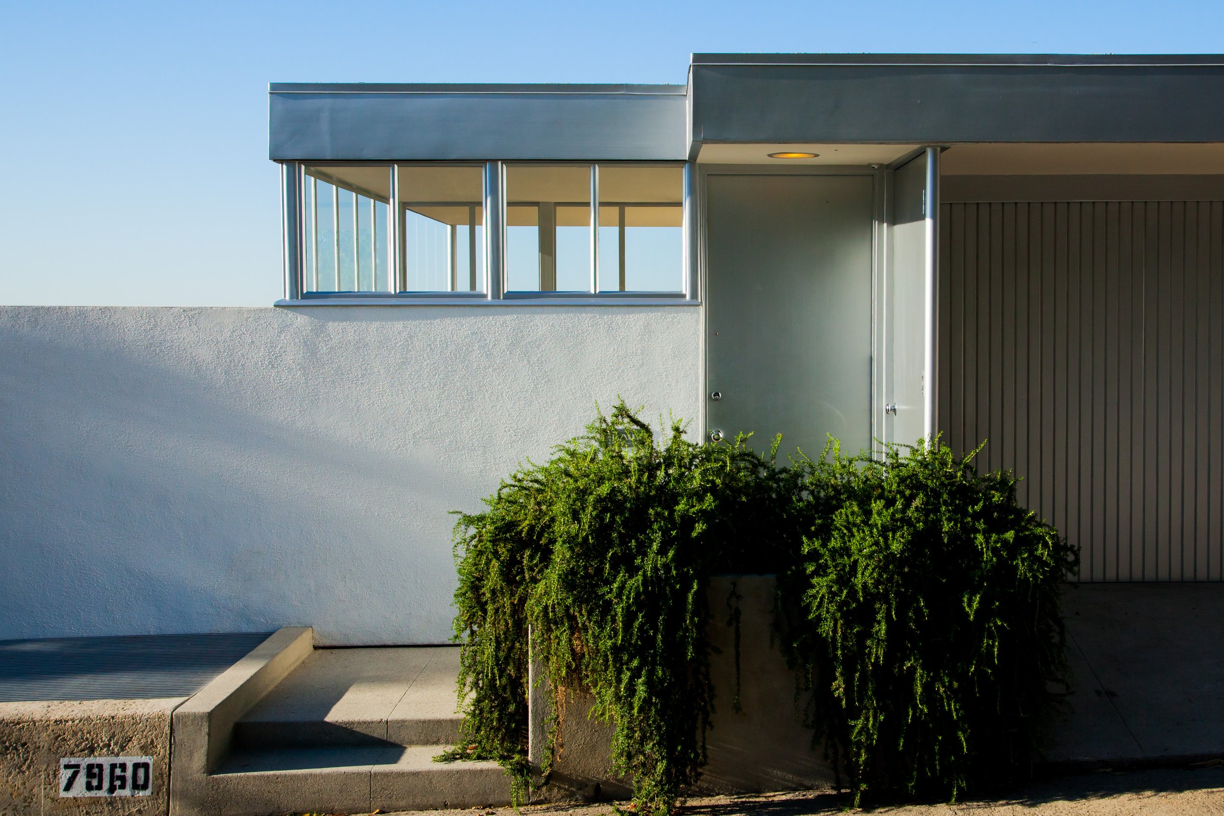 Modern mid-century home entrance with large windows, metal door, garage, and leafy shrubs