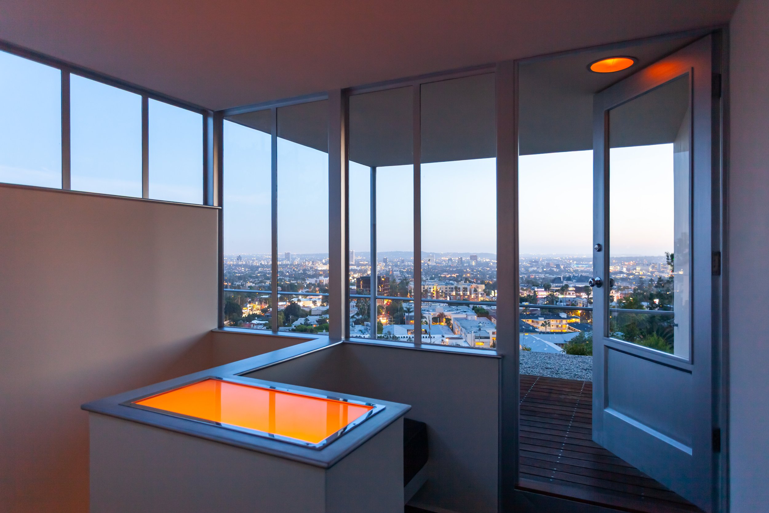 Modern home interior with illuminated square skylight, floor-to-ceiling windows, and a city view at dusk