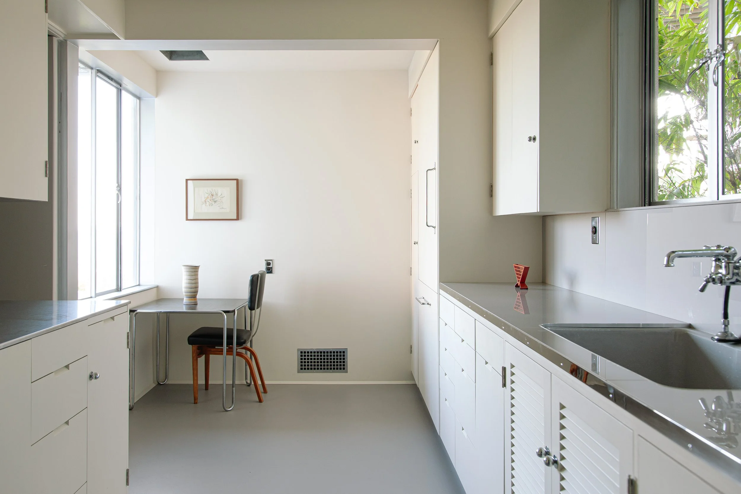 Bright modern white kitchen with gray countertops, large windows, custom cabinets, a dining table, and chairs