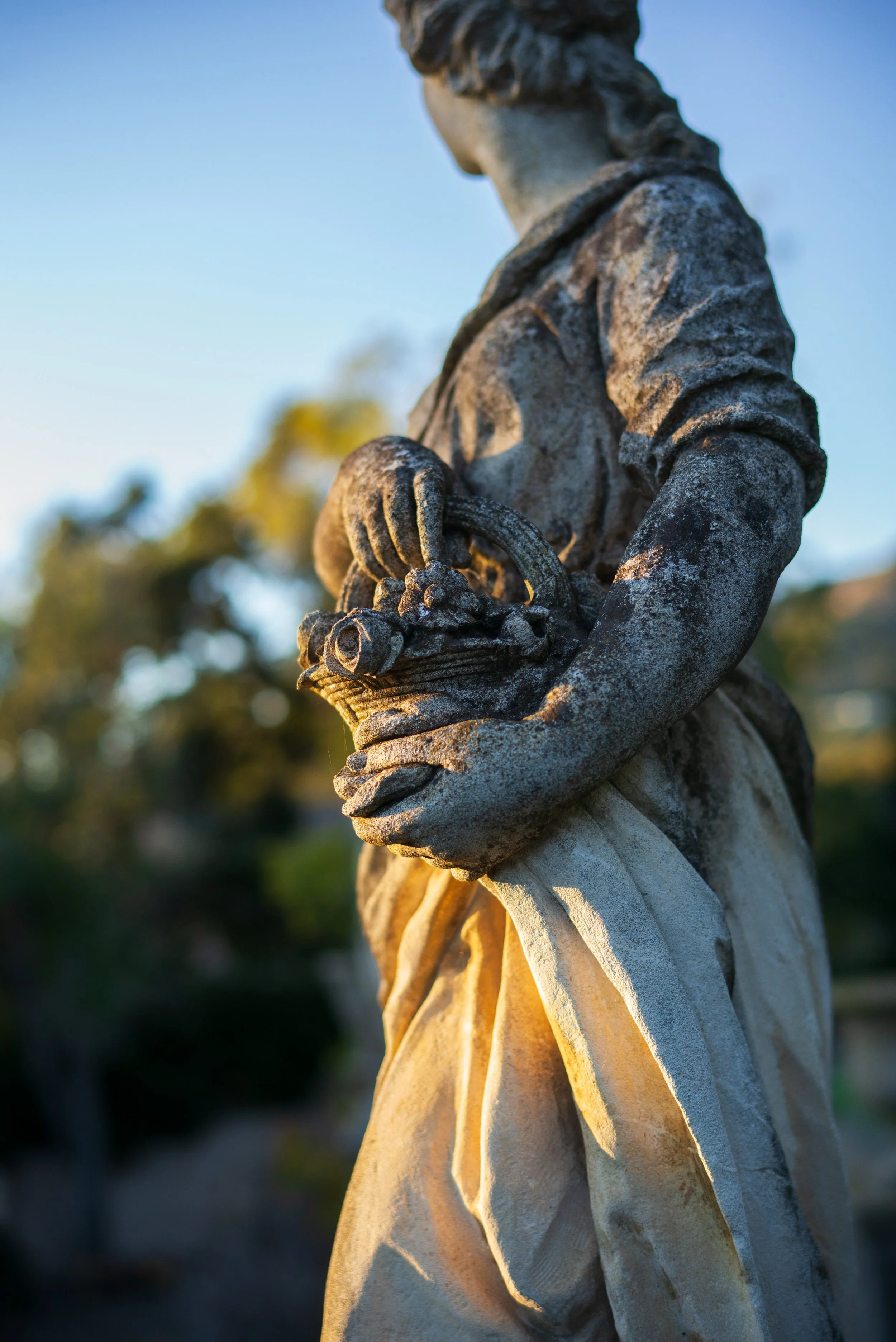 Close-up of a weathered stone statue of a robed figure holding a skull, illuminated by warm sunlight.