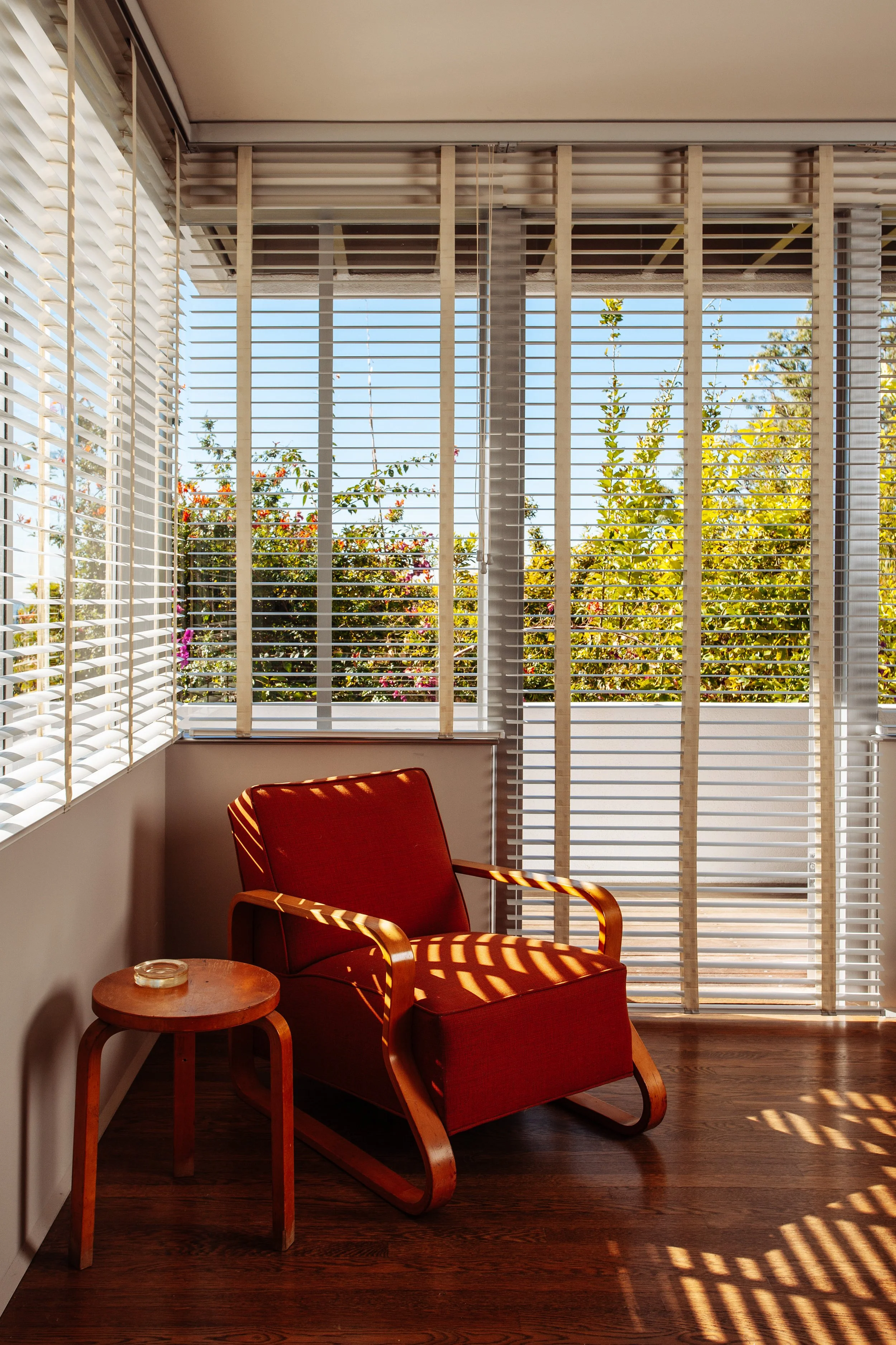 Sunlit red armchair and side table beside large windows with white blinds overlooking a leafy garden