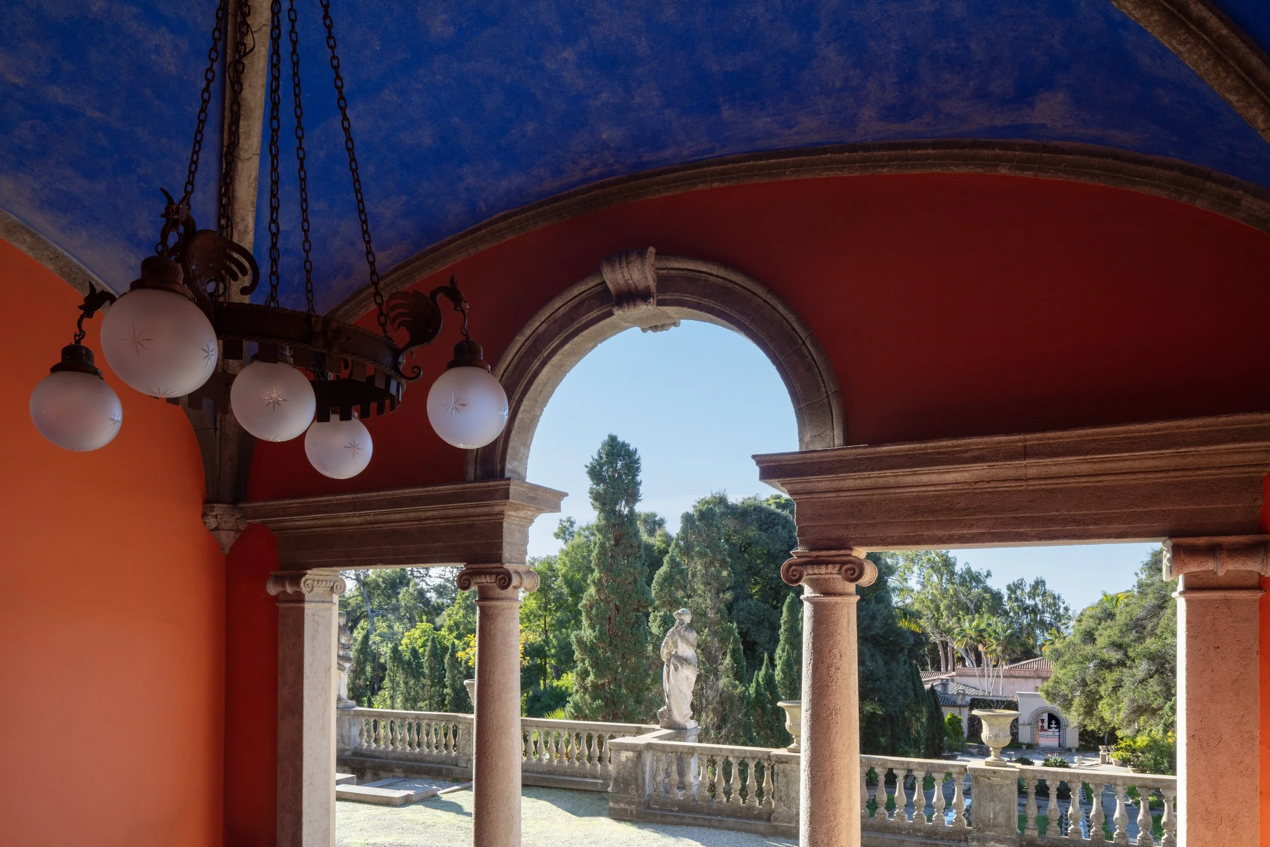 Ornate historic courtyard with red walls, blue vaulted ceiling, hanging globe chandelier, stone arches, columns, statues, and landscaped gardens