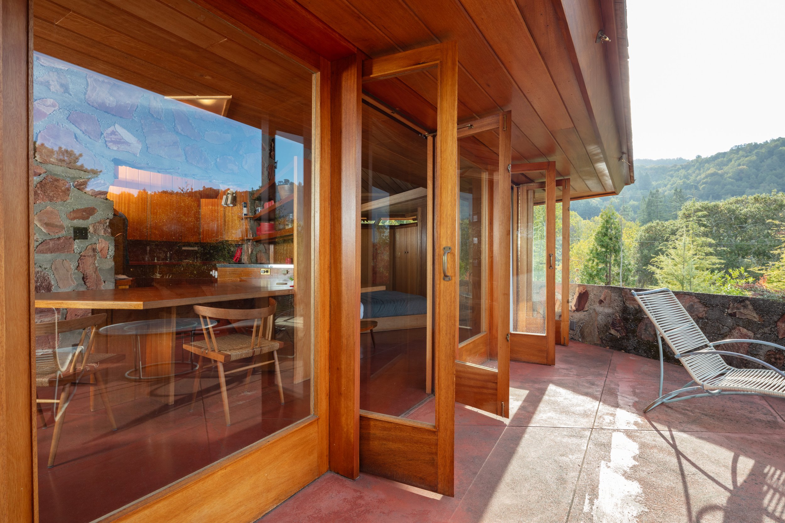 Wood-paneled modern home with open glass doors, stone fireplace, patio lounge chairs, and wooded mountain views